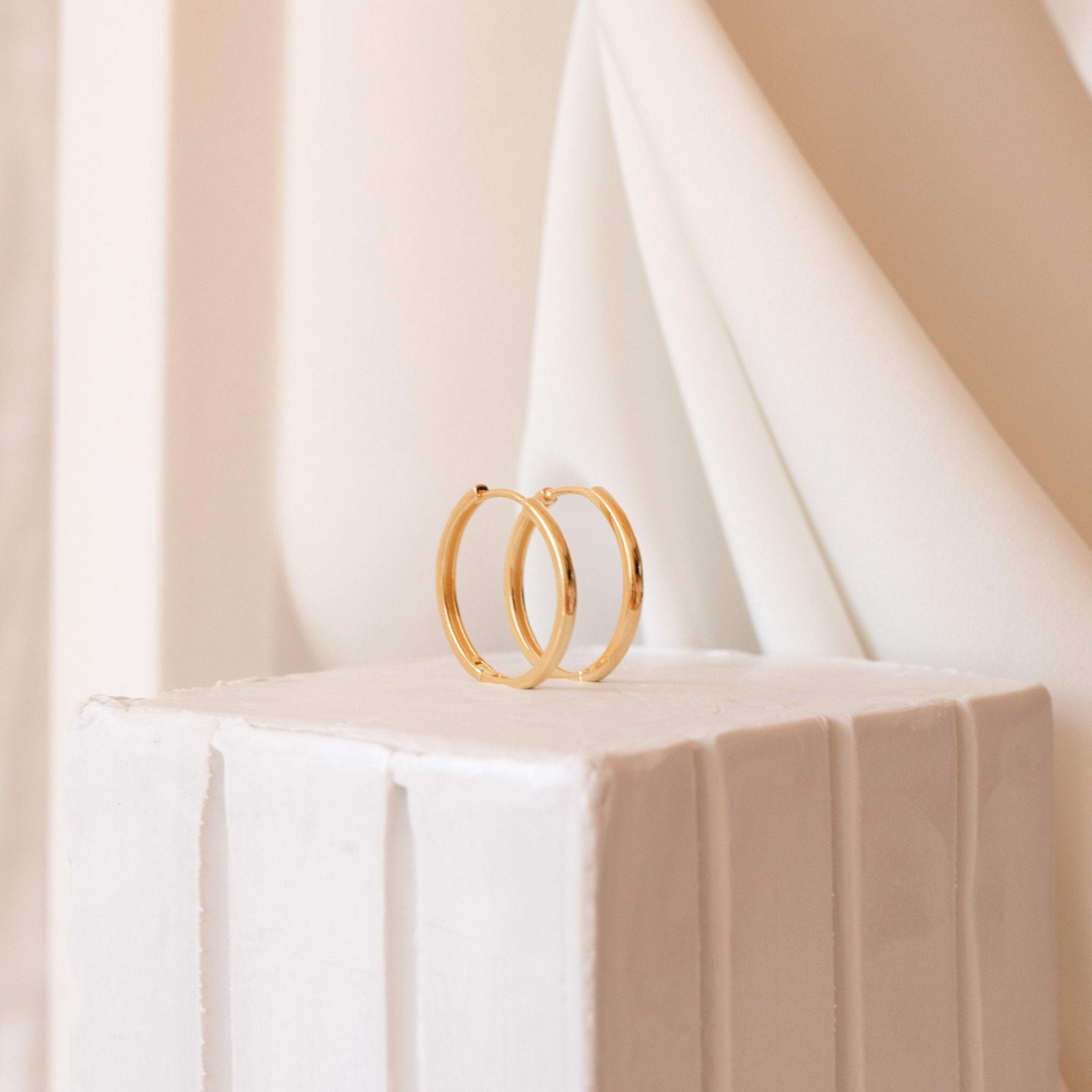 Classic Hoops are chic gold midi hoop earrings, featured on a white textured pedestal with a soft beige fabric background—an elegant choice for the modern city girl.