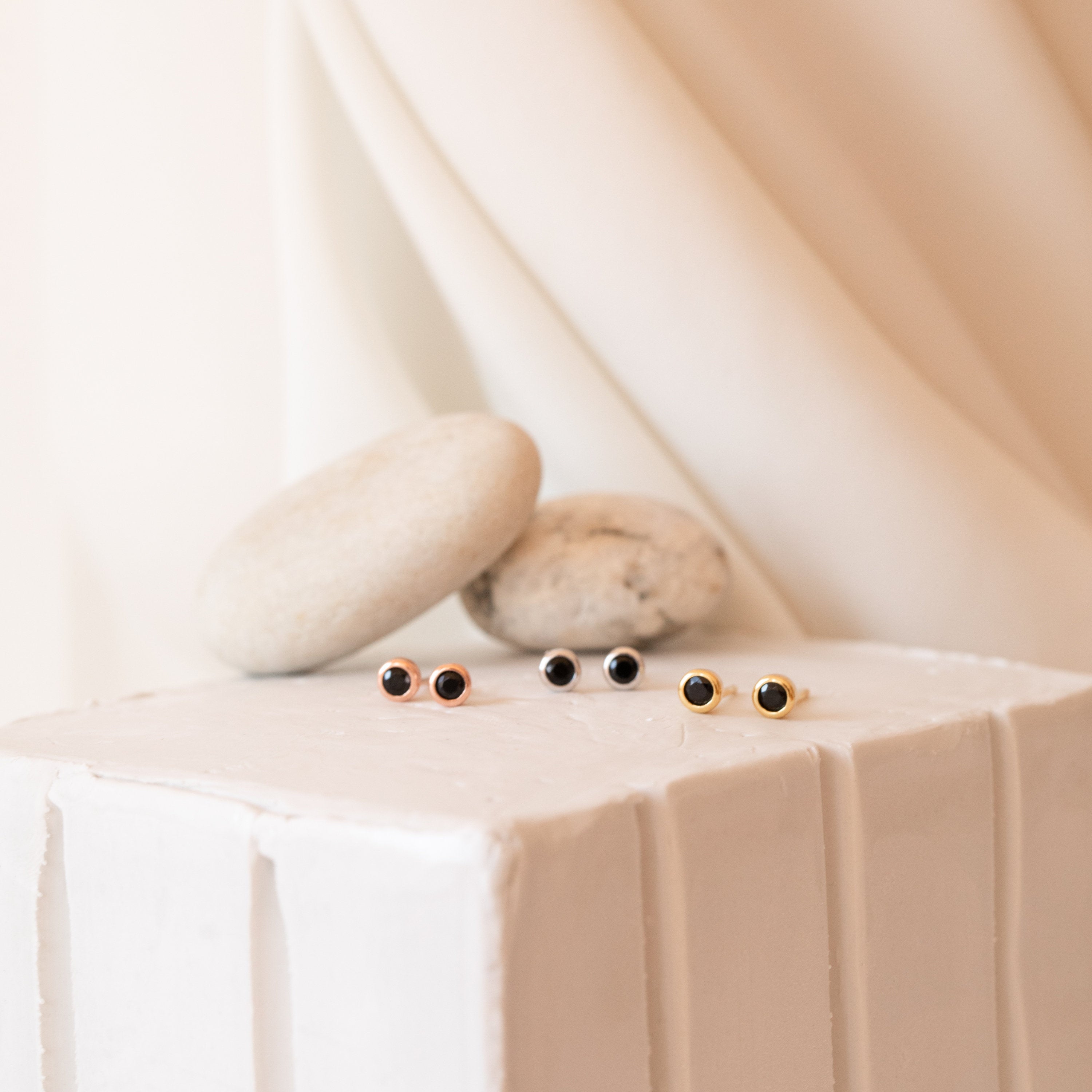 Three pairs of small stud earrings, featuring the Noir Diamond Studs, are displayed on a white textured surface with smooth stones in the background.