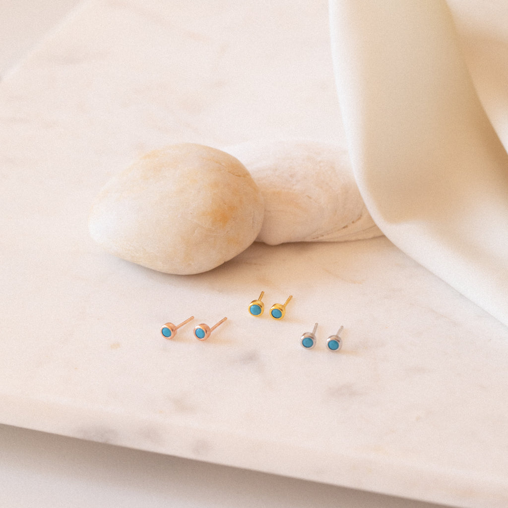 The Ariel Turquoise Studs, along with two other dainty earring pairs, are displayed on a marble surface accented by stones and ivory fabric in the background.