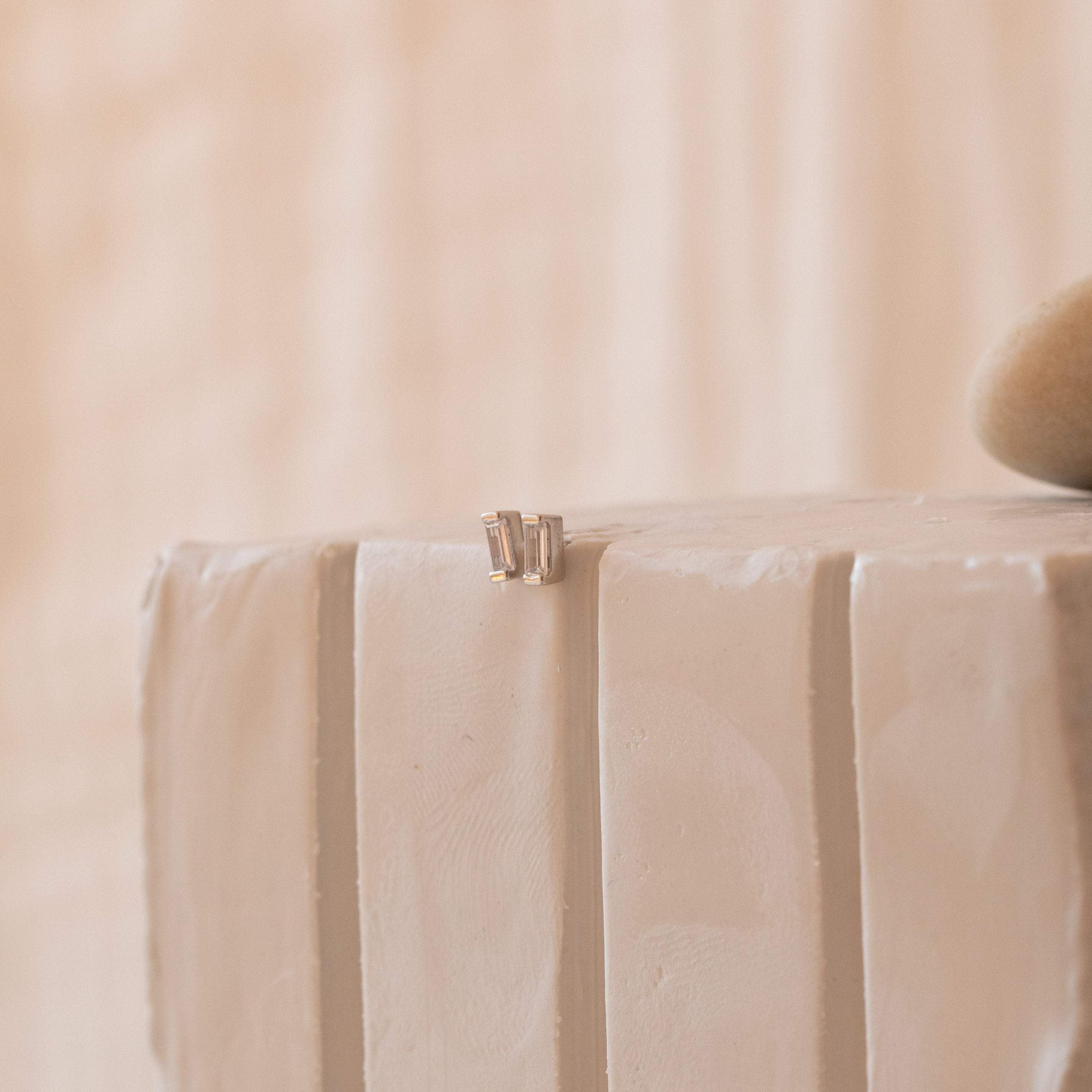 A pair of Baguette Diamond Studs, small and subtle, rest elegantly on the edge of a textured white surface.