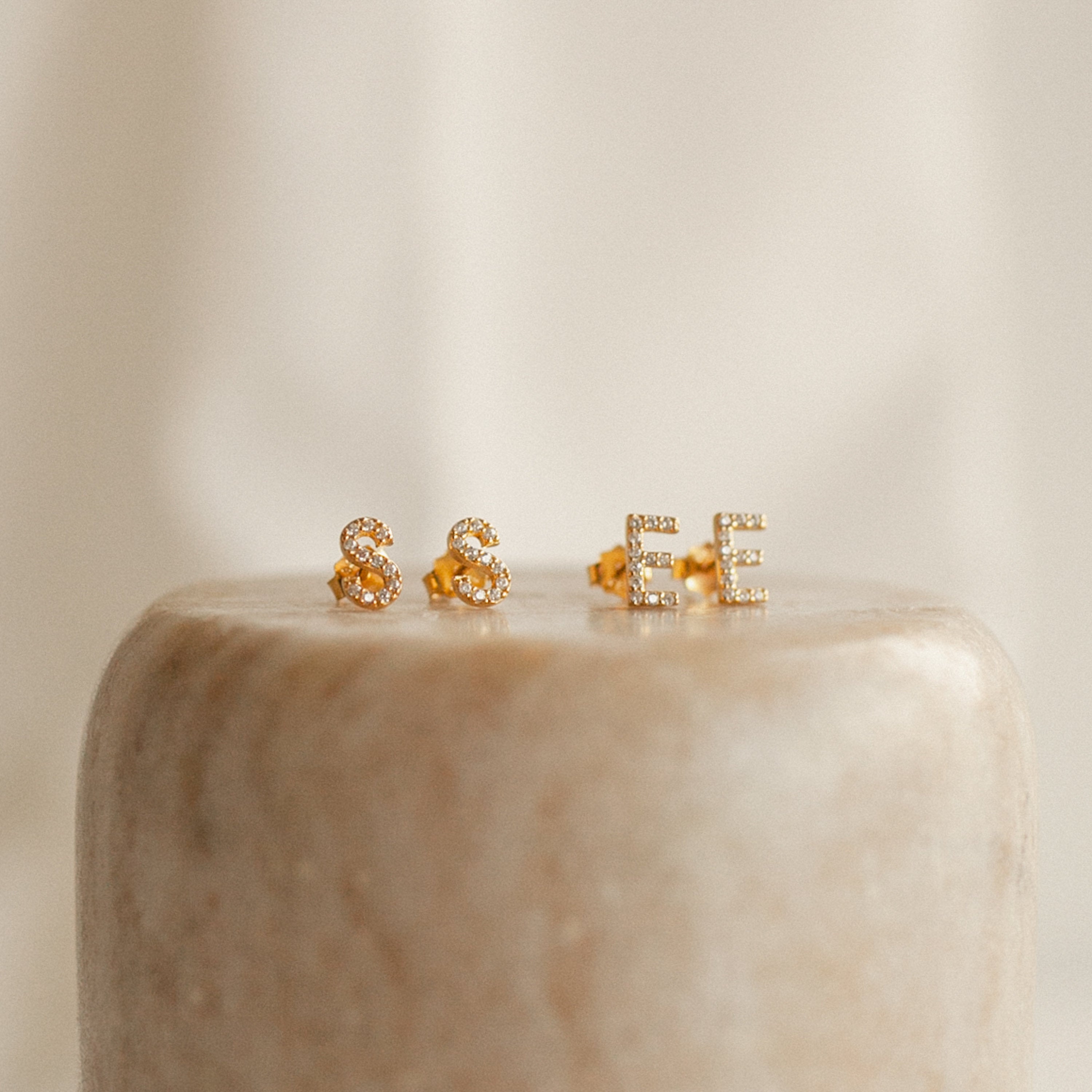 Pave Letter Studs shaped as S and E with sparkling crystals, shown on a beige rounded surface—perfect personalized earrings for a minimal style.
