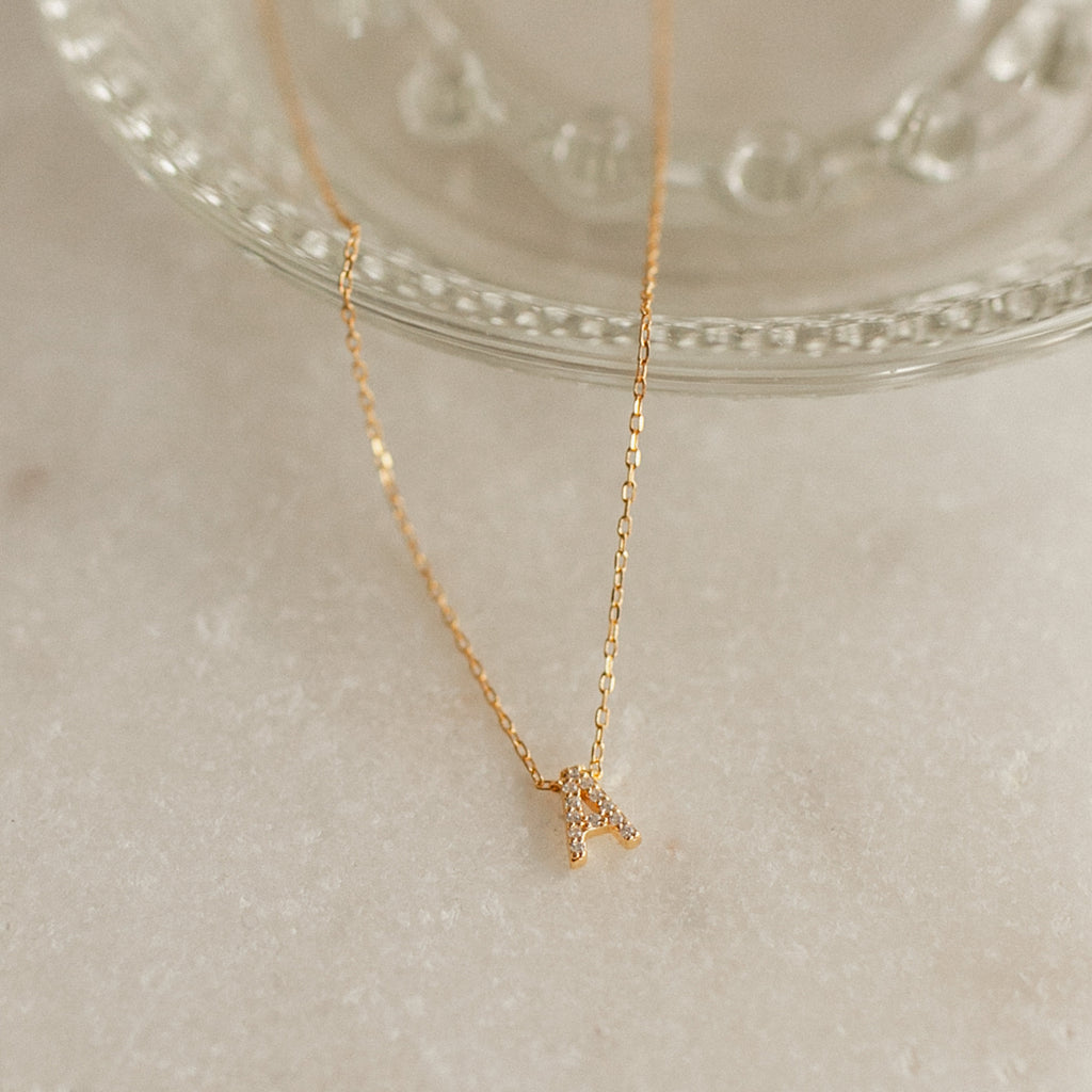 The Pave Initial Necklace features a small letter A pendant set with sparkling pave stones, displayed on a light, marble-like surface—an elegant and personalized jewelry piece.