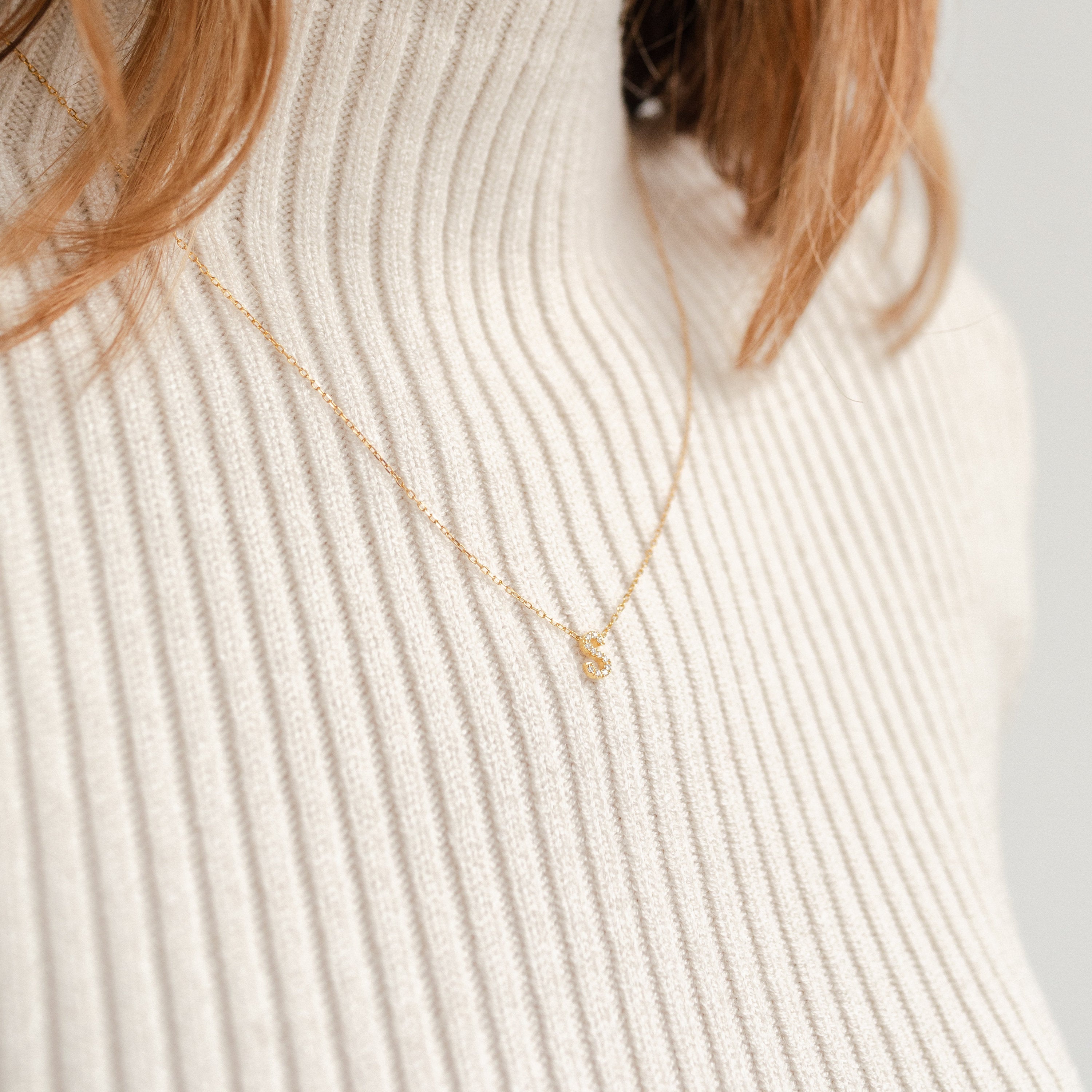 The Pave Initial Necklace in Rose Gold, featuring a delicate pendant, adds a personalized touch when elegantly styled over a cream ribbed turtleneck sweater.
