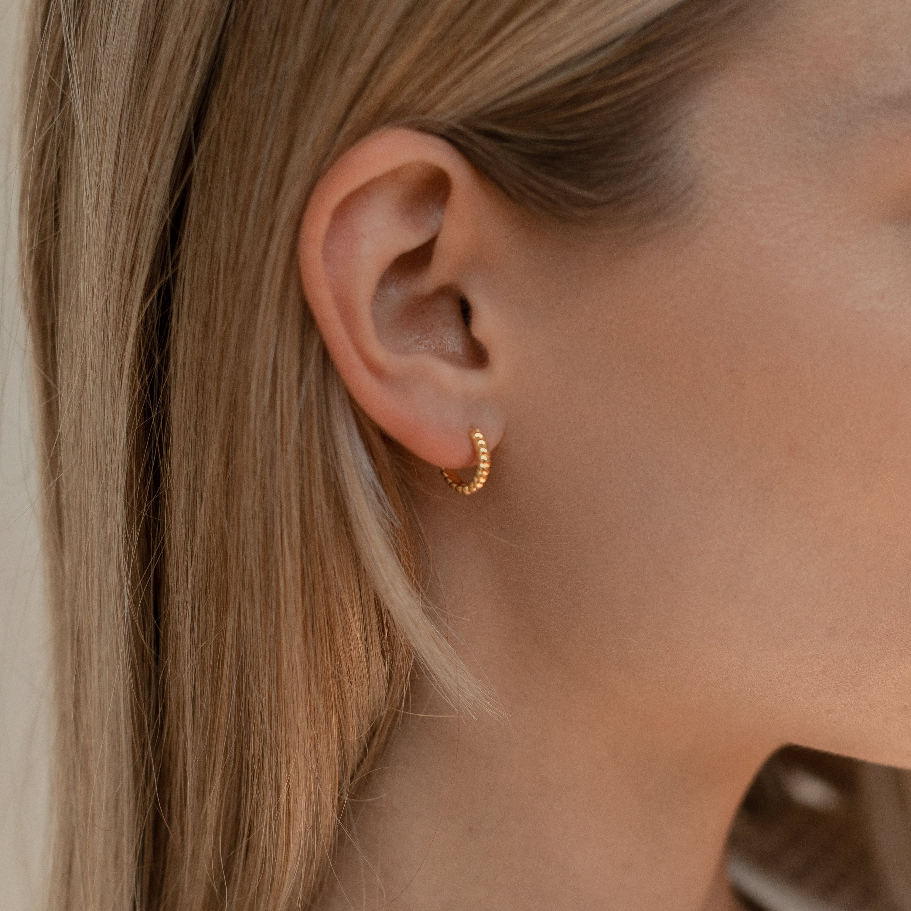 Close-up of a woman's ear with straight blonde hair, wearing Thea Twisted Huggies in 18K Gold.