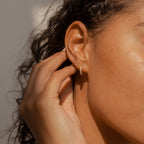 A close-up of a person touching their ear, wearing Vera Pave Huggies in Rose Gold—elegant minimalist earrings ideal for daily wear—in warm lighting.