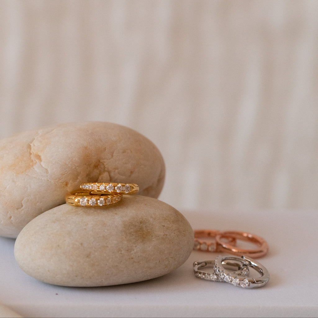 The Vera Pave Huggies in Rose Gold, displayed on smooth beige stones with minimalist rings and earrings, create a refined everyday jewelry collection against a soft background.