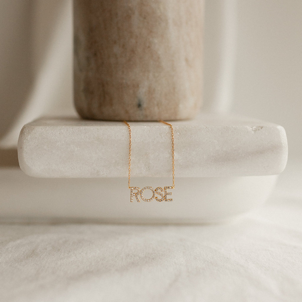 The Pave Name Necklace in Rose Gold, personalized with block letters spelling “ROSE,” is displayed on a white marble surface under soft, neutral lighting.