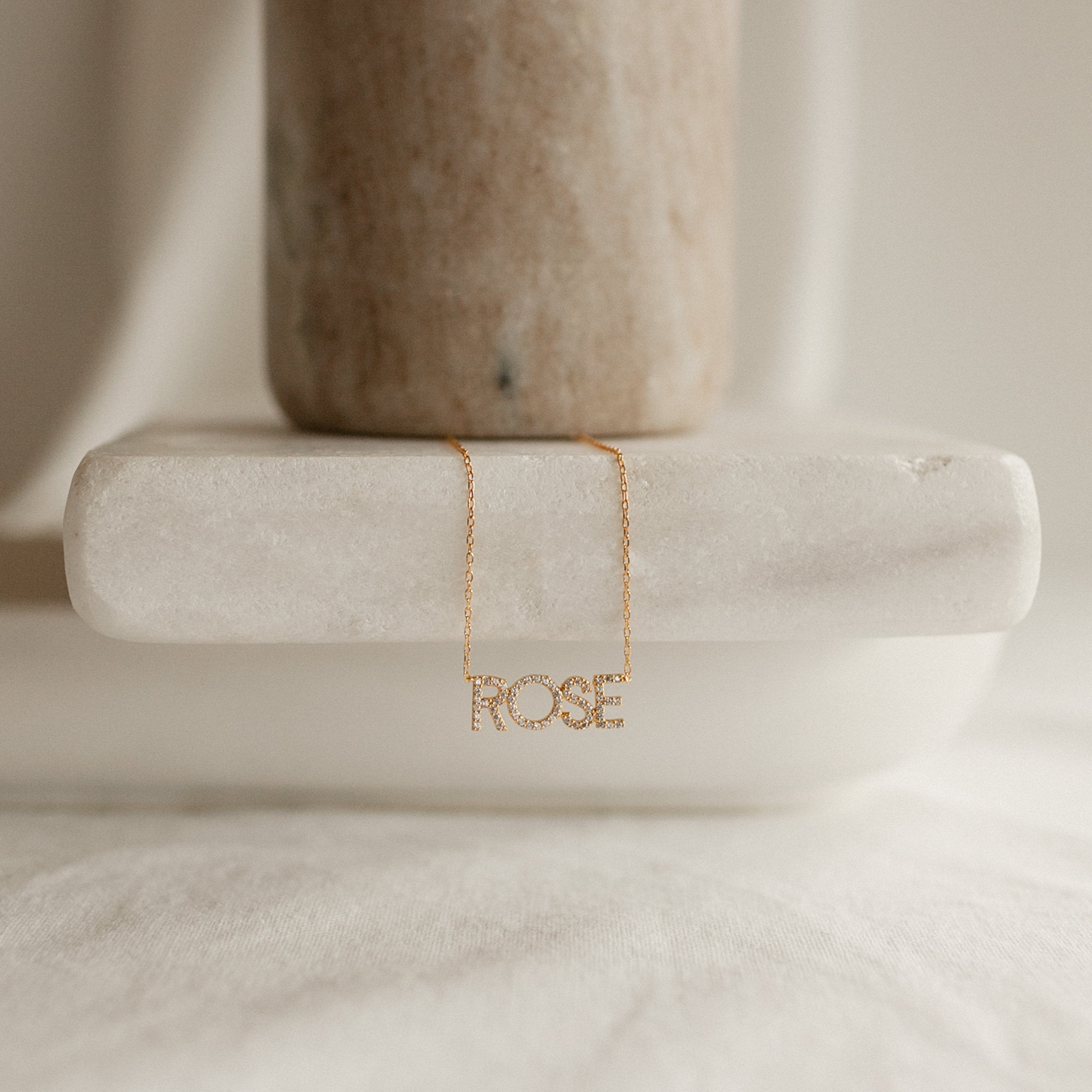 The Pave Name Necklace in Rose Gold, personalized with block letters spelling “ROSE,” is displayed on a white marble surface under soft, neutral lighting.