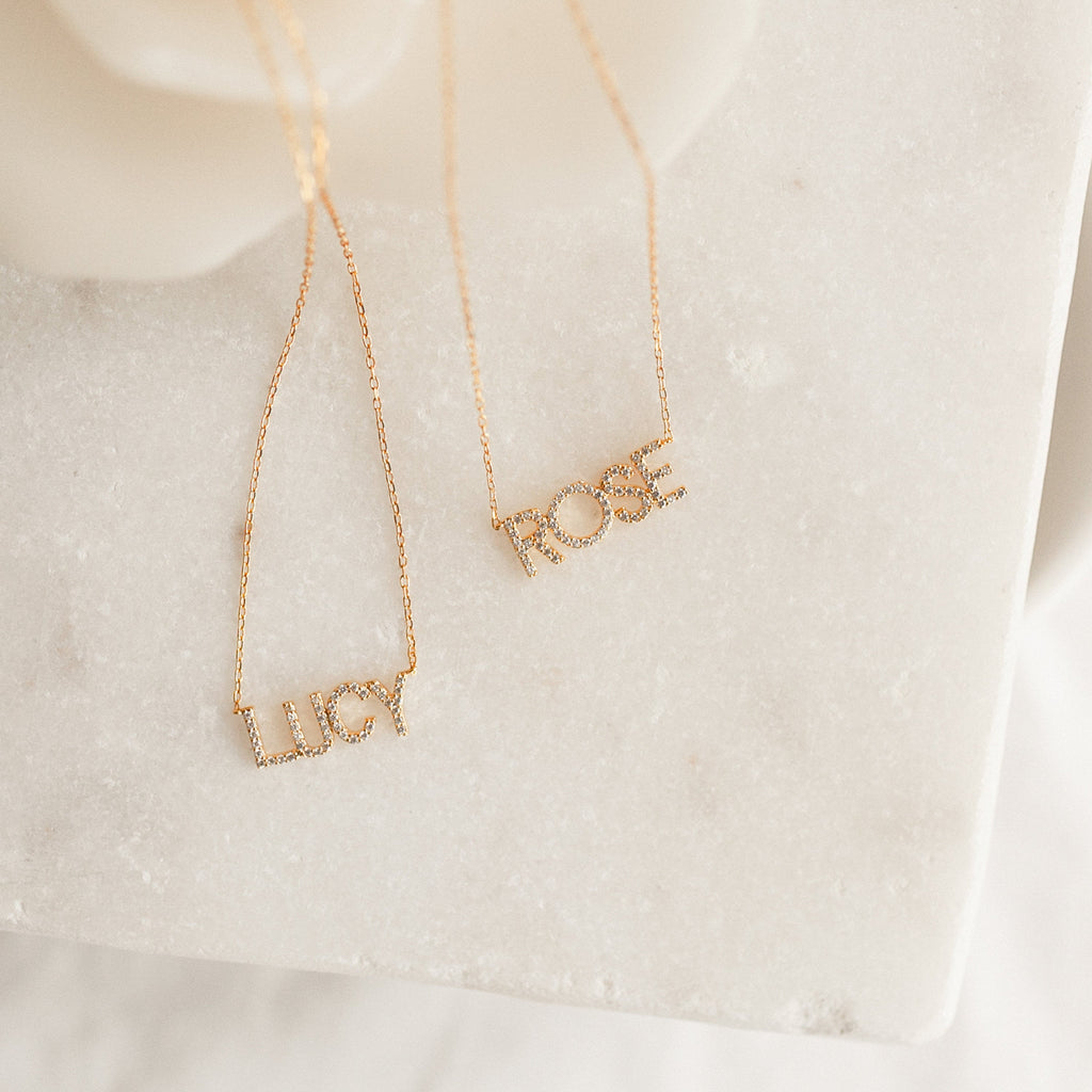 Two Pave Name Necklaces with personalized block letters—LUCY and ROSE—are displayed as gold name pendants on a white marble surface for a timeless look.