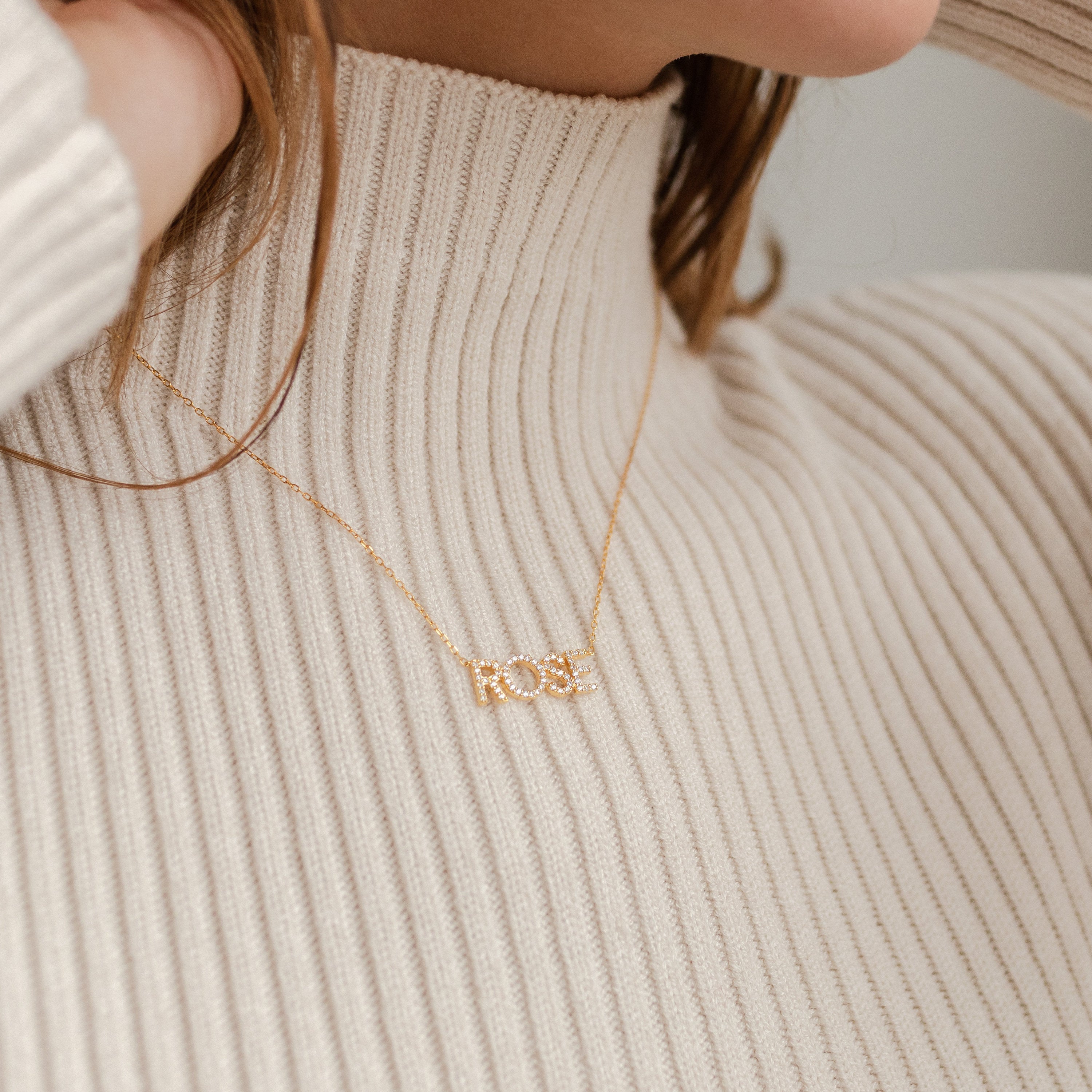 Close-up of a woman wearing a beige ribbed turtleneck and a rose gold Pave Name Necklace with personalized block letters spelling ROSE.