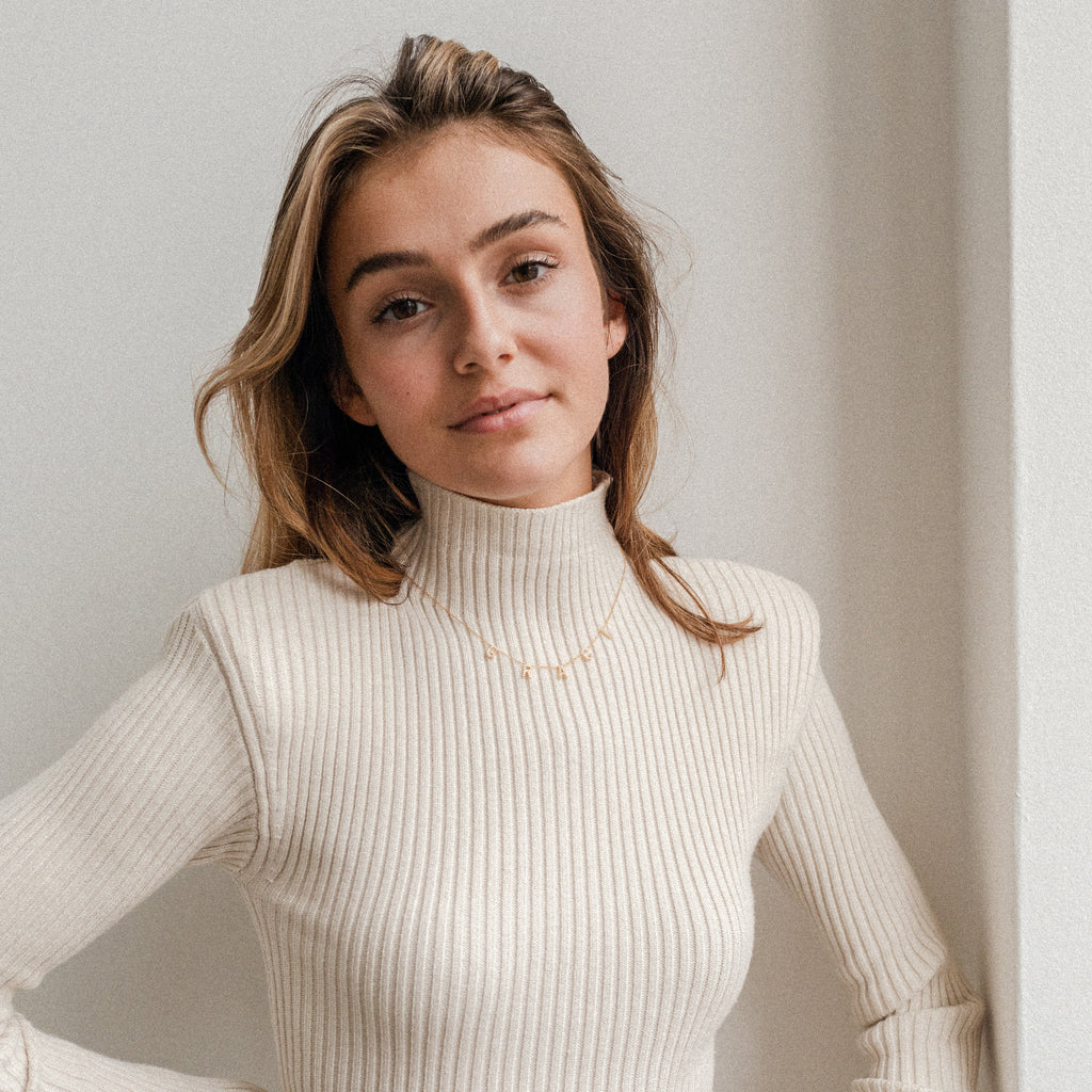 A woman with long hair wears a cream ribbed turtleneck sweater and a Pave Letter Necklace, standing against a white wall and smiling softly.