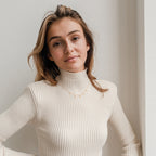 A woman with long hair wears a cream ribbed turtleneck sweater and a Pave Letter Necklace, standing against a white wall and smiling softly.