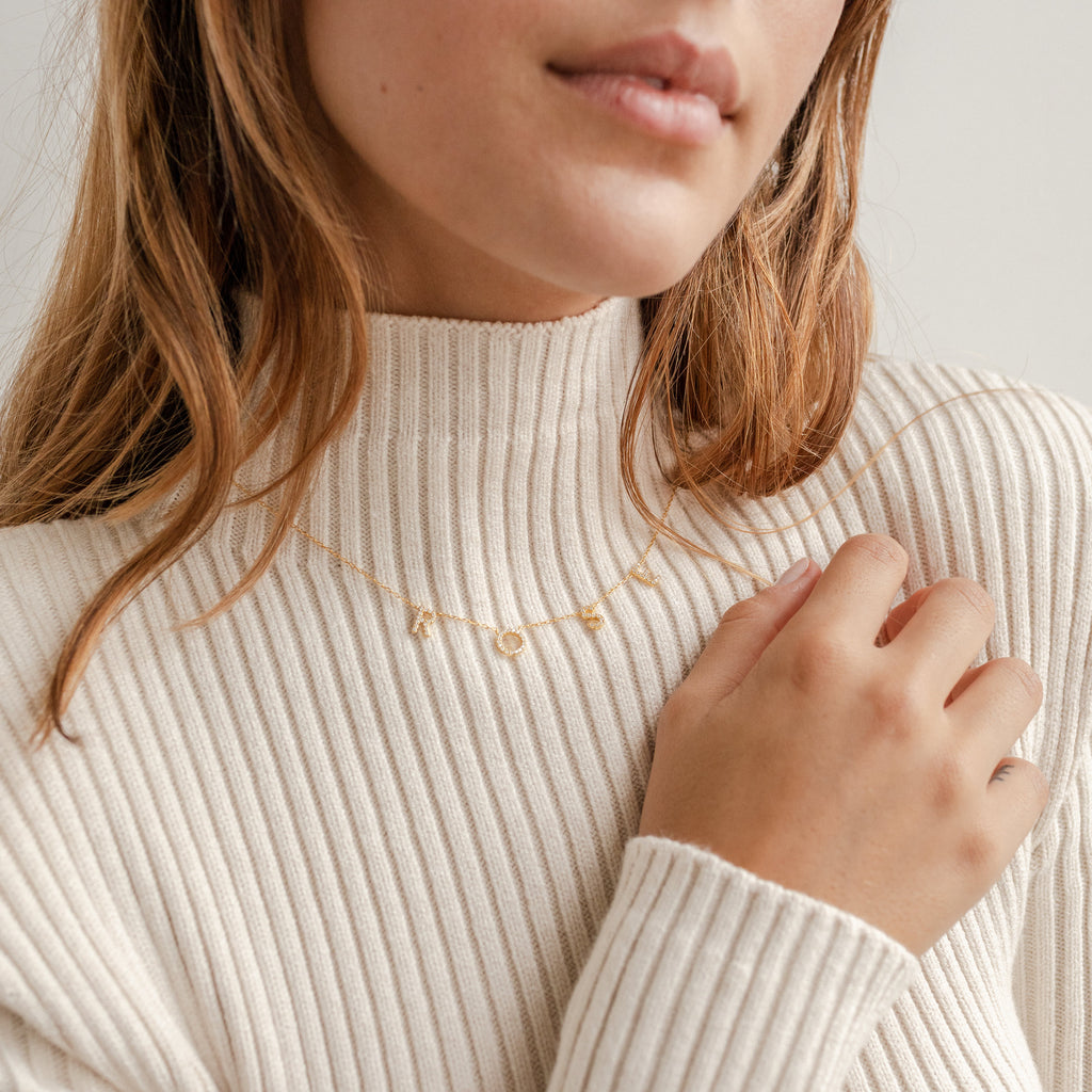 A woman in a cream ribbed sweater holds a custom Pave Letter Necklace in Rose Gold, featuring charms that spell ROSY—an ideal gift for fans of personalized jewelry.