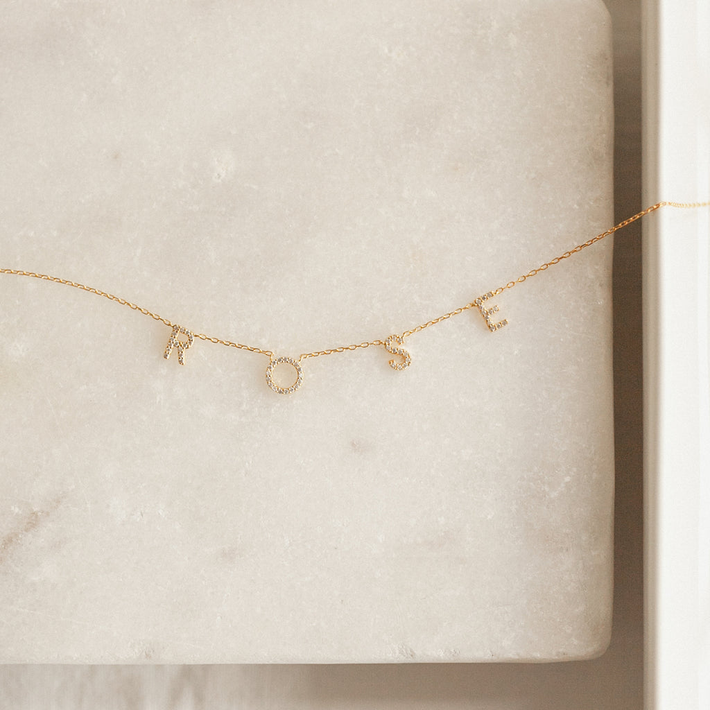 The Pave Letter Necklace features delicate letter charms spelling ROSE on a light marble surface, offering a customizable and stunning piece of personalized jewelry.