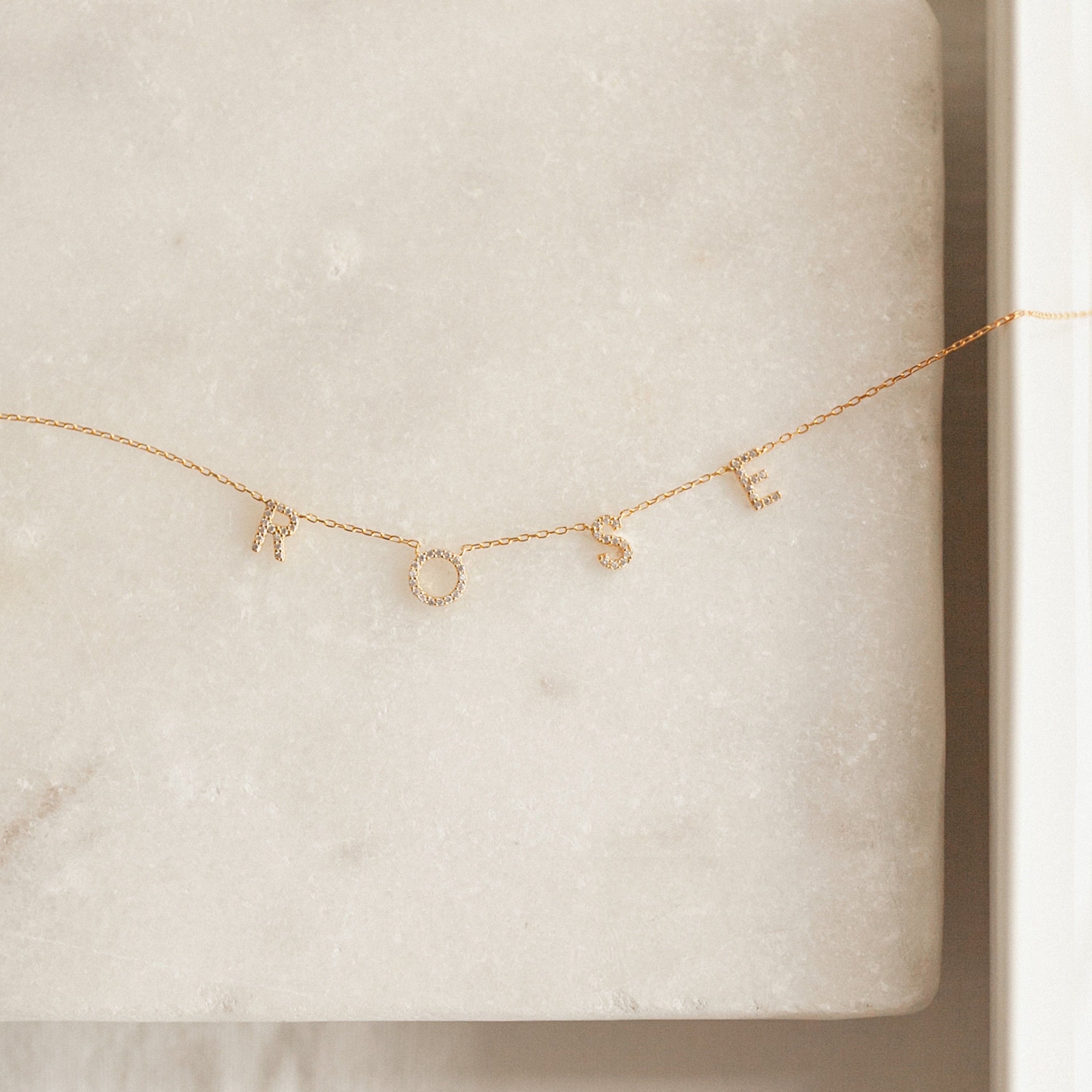 The Pave Letter Necklace features delicate letter charms spelling ROSE on a light marble surface, offering a customizable and stunning piece of personalized jewelry.