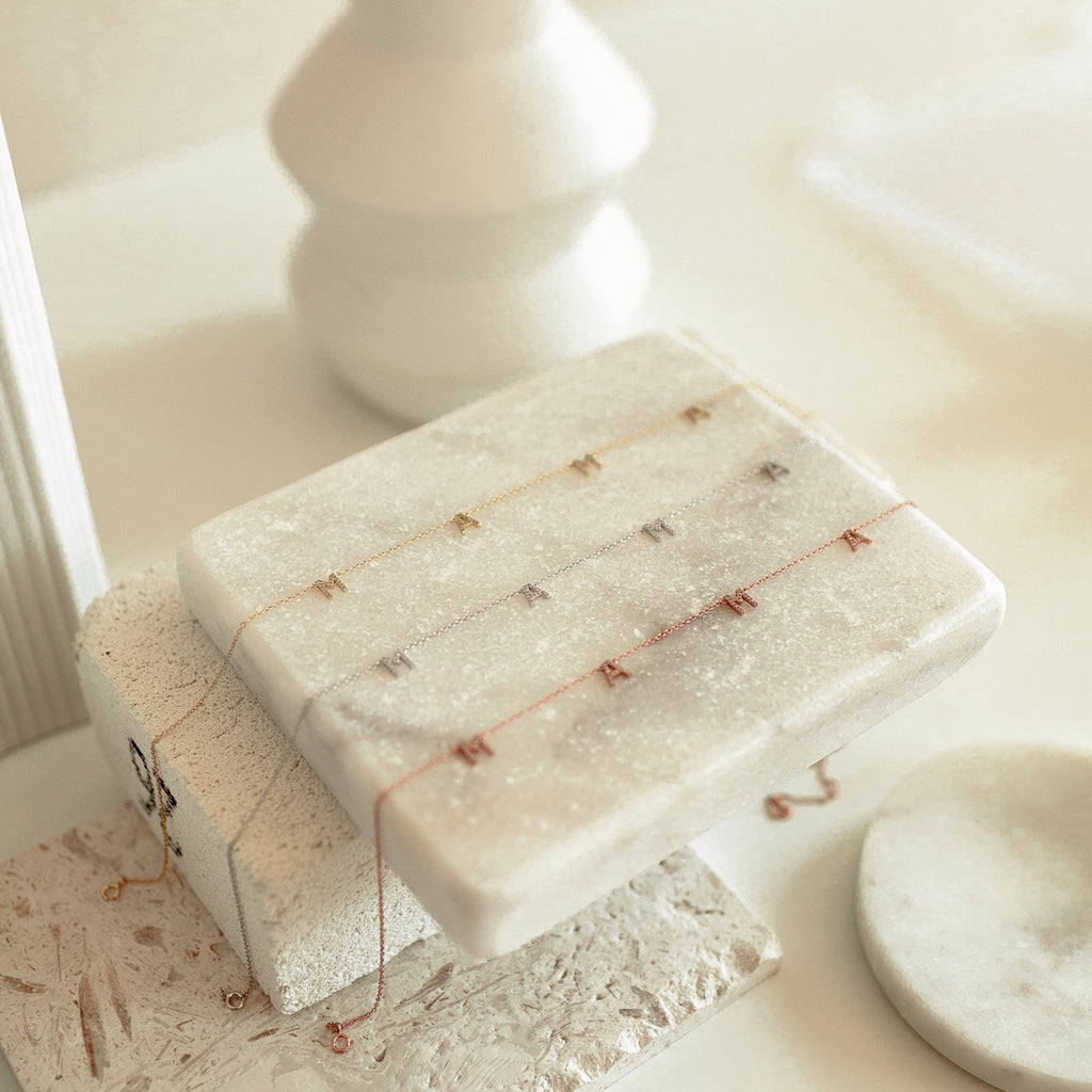The Pave Mama Letter Necklace, available in gold, silver, and rose gold, shines on a white marble stand with soft lighting—perfect as a motherhood accessory and a thoughtful gift for mom.