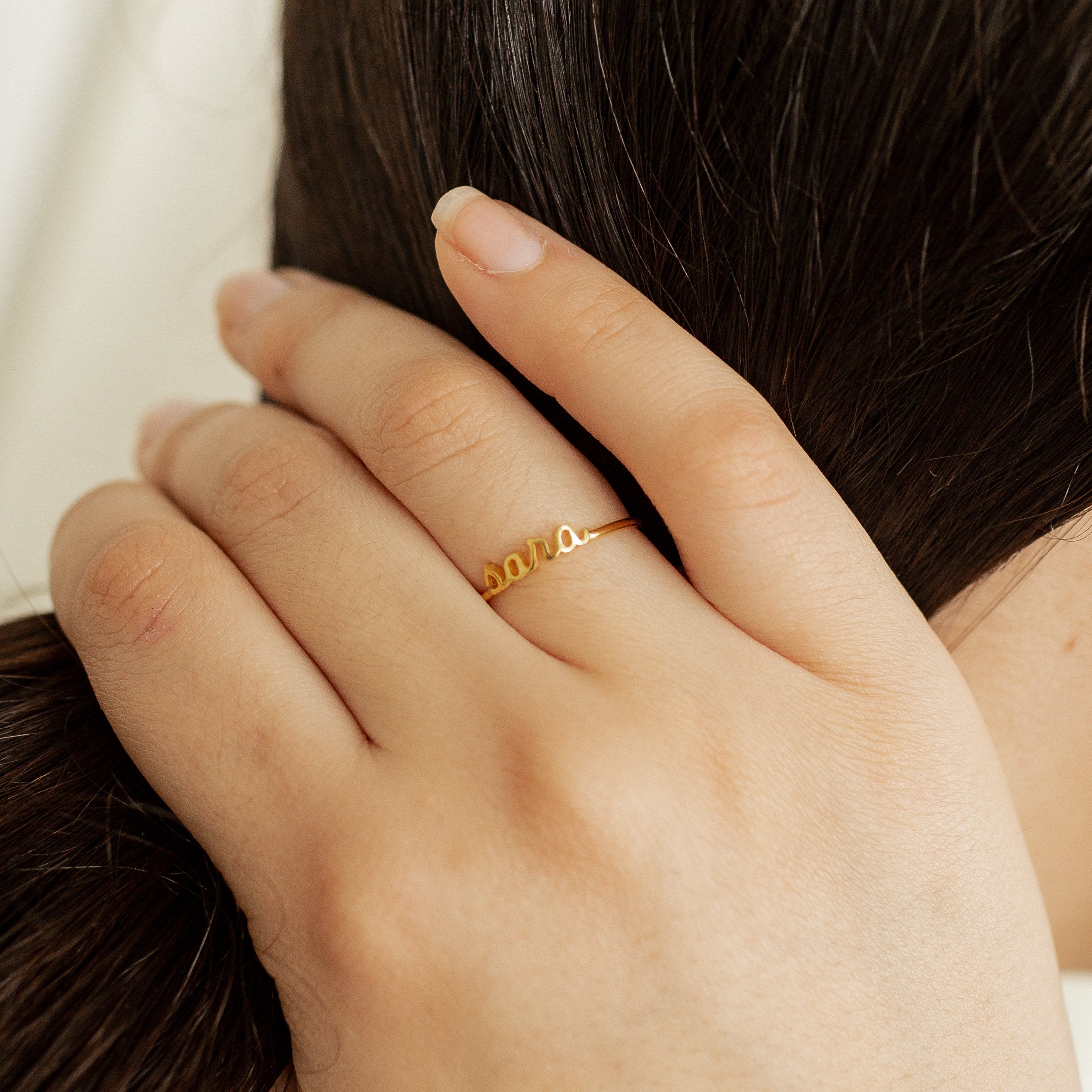A hand wearing the Petite Tiny Name Ring spelling "sara" rests on long, dark brown hair.