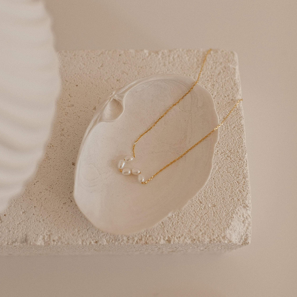 Gold chain necklace with five small oval freshwater pearls arranged in a soft curve Necklace displayed on a white ceramic dish on a textured surface.
