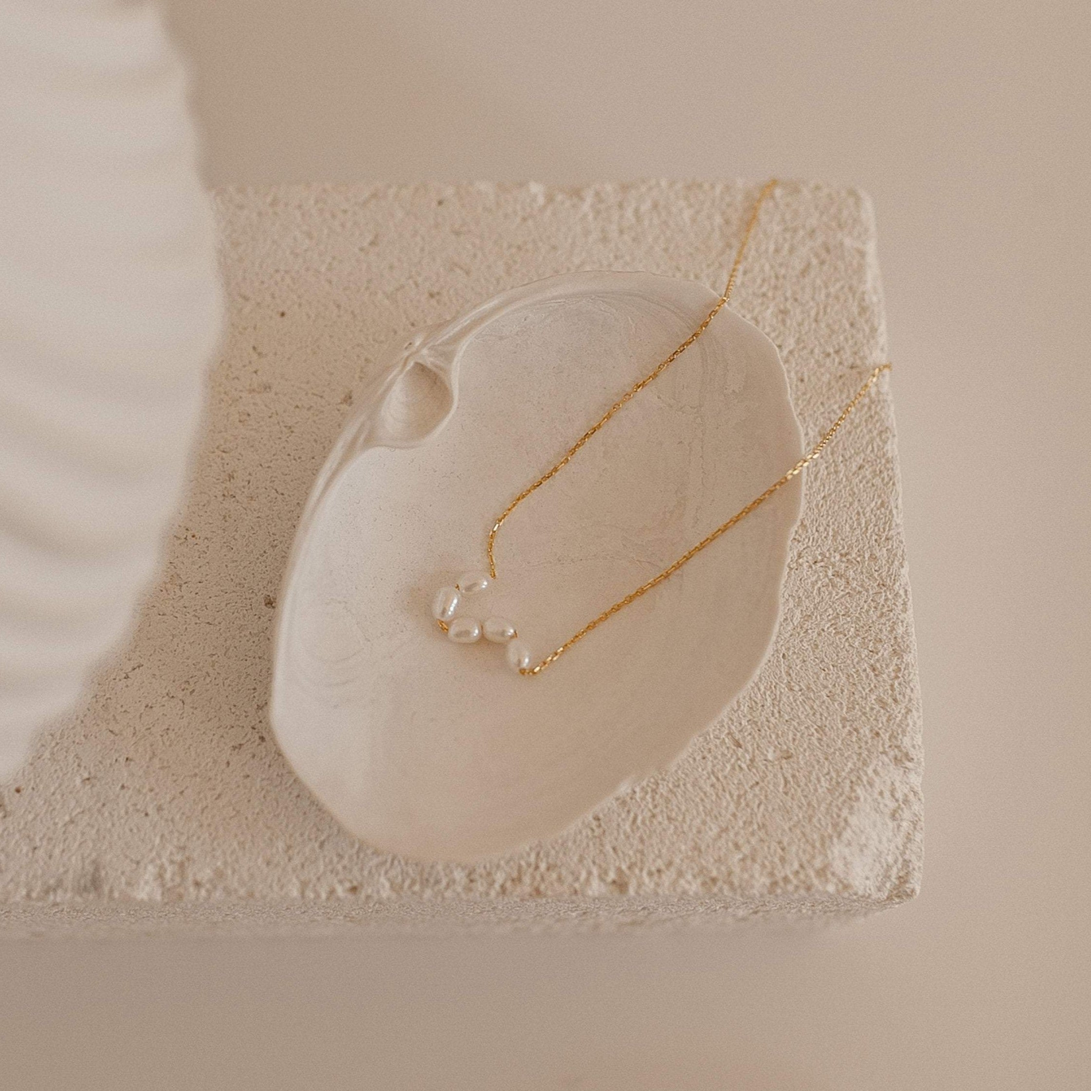 Gold chain necklace with five small oval freshwater pearls arranged in a soft curve Necklace displayed on a white ceramic dish on a textured surface.