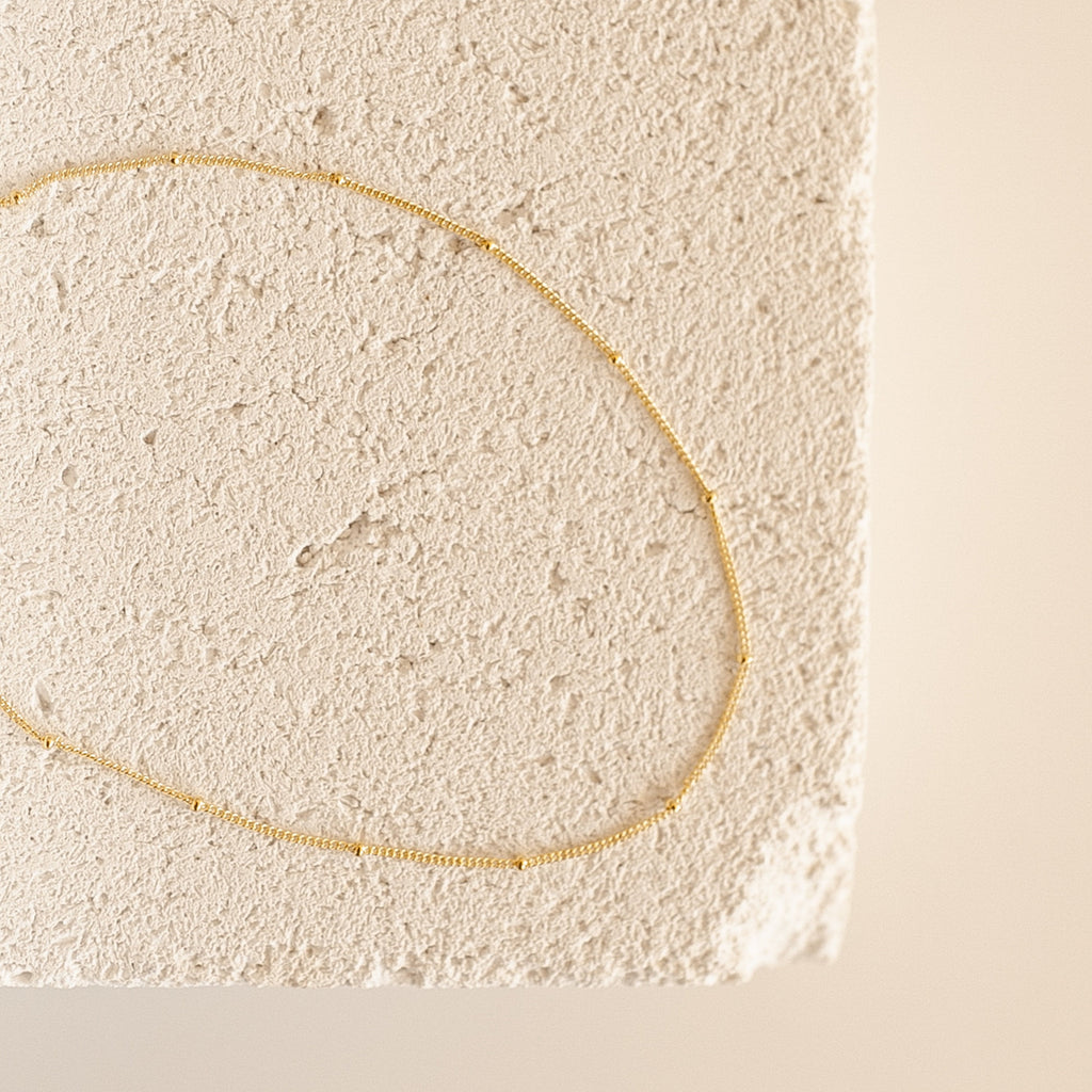 A gold Satellite Necklace arranged in an oval shape atop a textured, off-white stone surface.