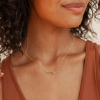 Close up of a woman wearing a delicate gold necklace featuring two interlocking oval links, styled simply against a rust colored tank top.