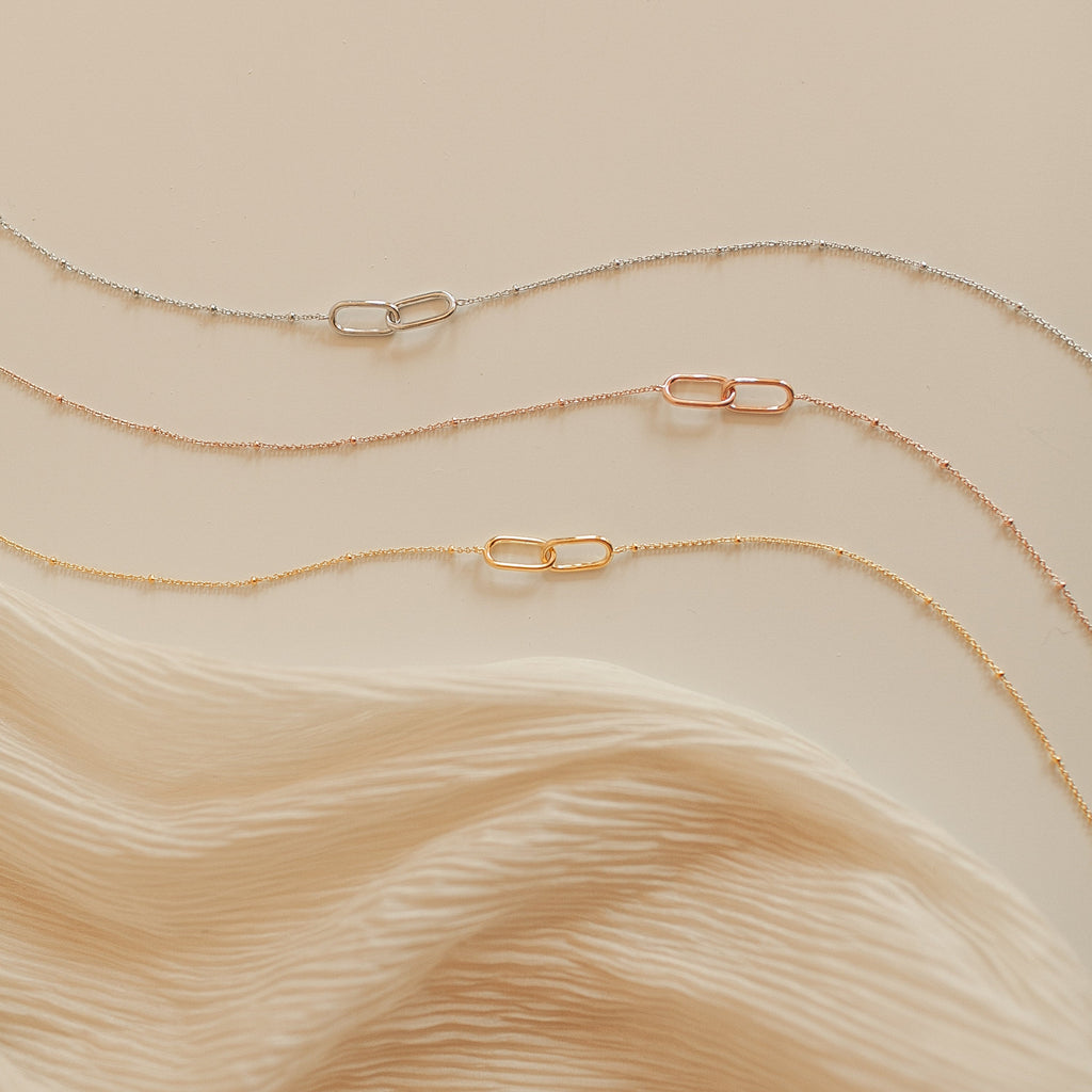 The Linked Pendant Necklace in Rose Gold is featured, displayed on soft, cream-colored fabric.