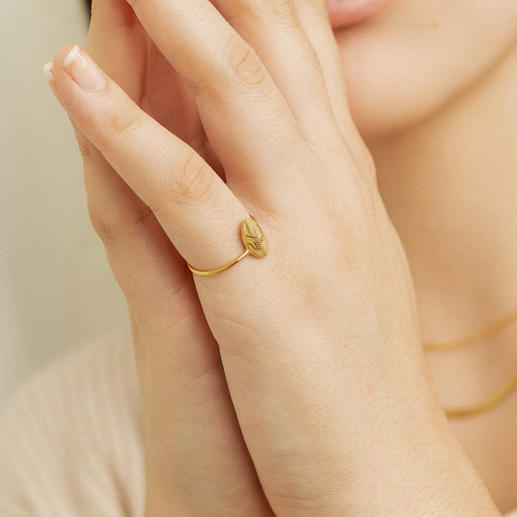 A hand adorned with the Gestures of Love Ring, featuring an engraved design, rests near a person’s face and is paired with a gold necklace—figurative jewelry expressing love through symbolic hand gestures.
