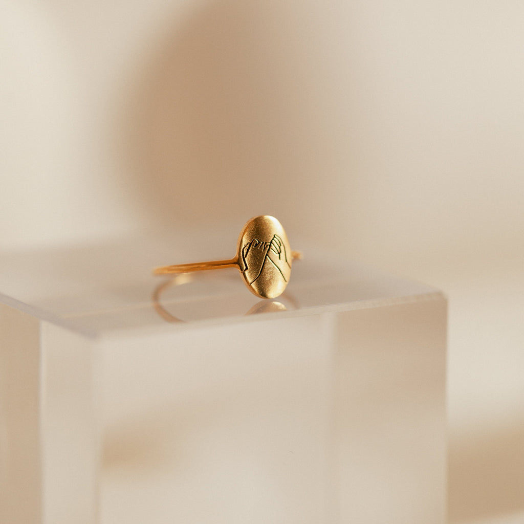 The Gestures of Love Ring, featuring an engraved hand design, is shown on a clear frosted stand—a stunning piece of figurative jewelry that captures meaningful hand gestures.