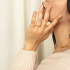 A woman wearing a gold necklace and the Pave Initial Ring gently touches her chin with her hand.