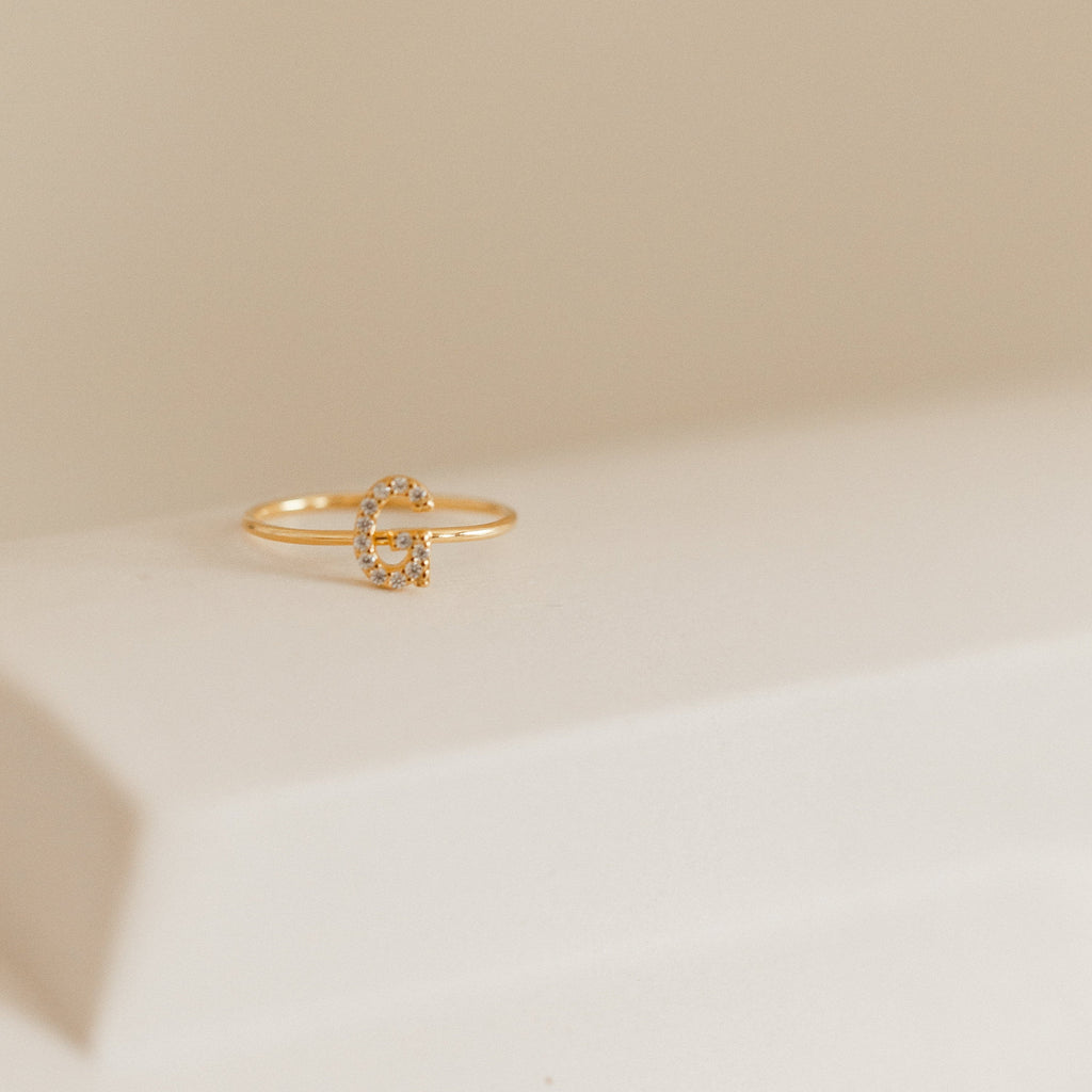 The Pave Initial Ring, featuring a diamond-encrusted gold letter G, rests on a white surface with a beige background.