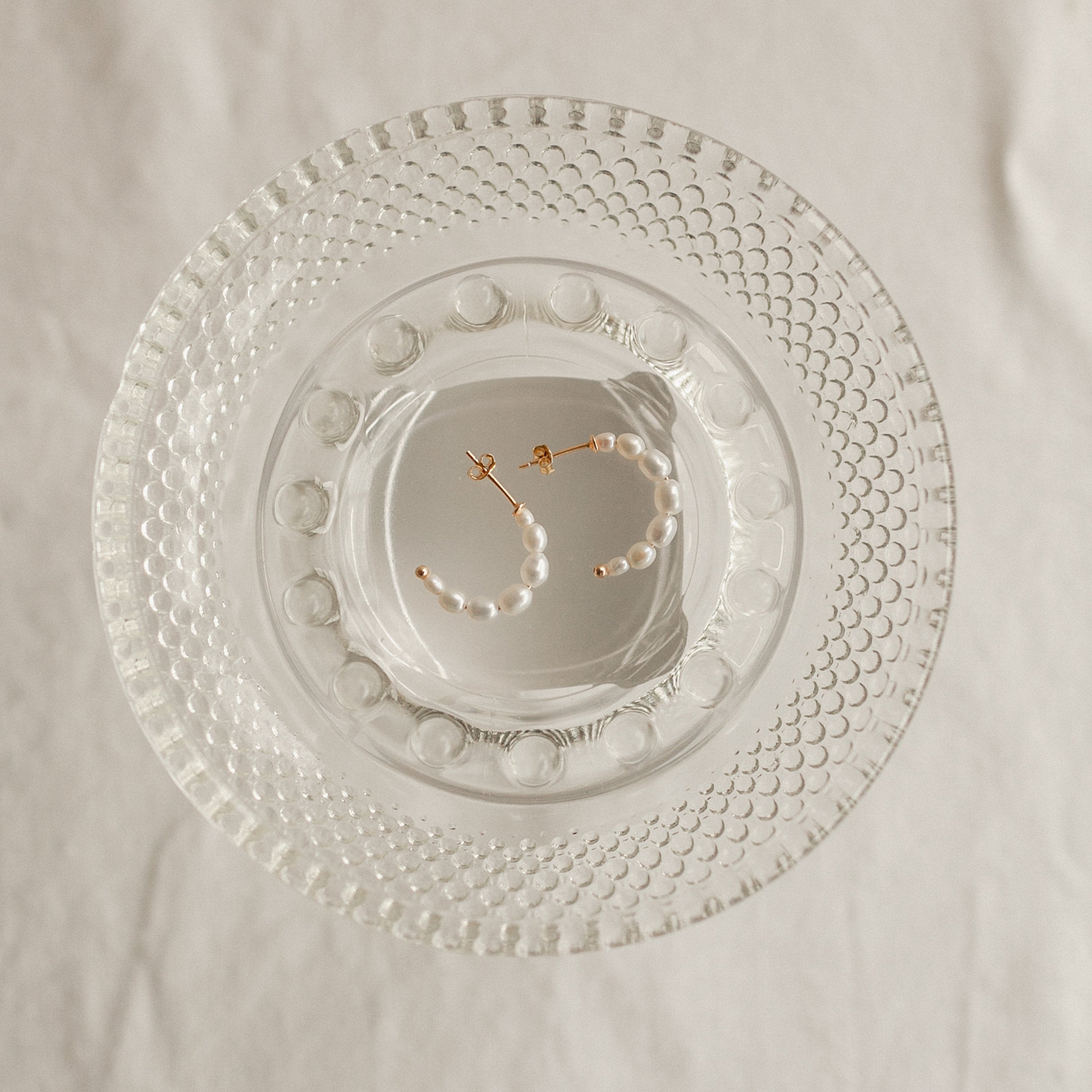 The Pearl Hoops feature white pearls and rest on a textured clear glass plate, making these earrings an elegant accessory for everyday wear.