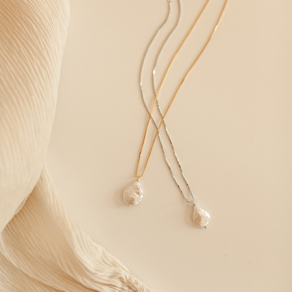 Two Sadie Pearl Necklaces with freshwater pearl pendants—one on a gold chain, one on a silver chain—rest on a cream fabric background, making an elegant bridesmaids gift.