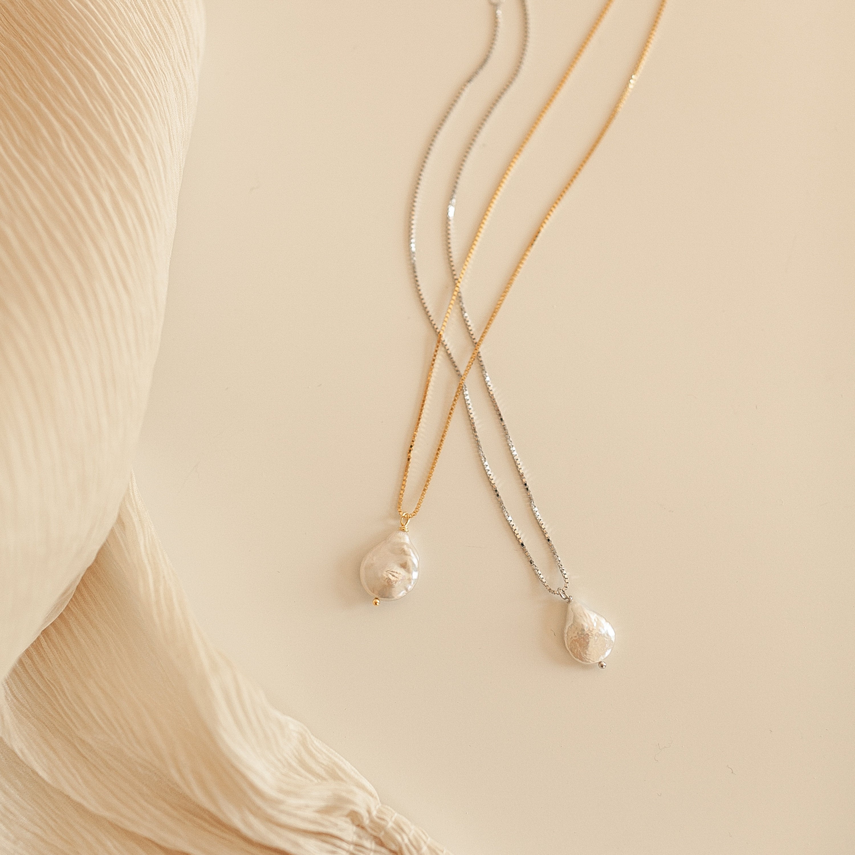 Two Sadie Pearl Necklaces with freshwater pearl pendants—one on a gold chain, one on a silver chain—rest on a cream fabric background, making an elegant bridesmaids gift.