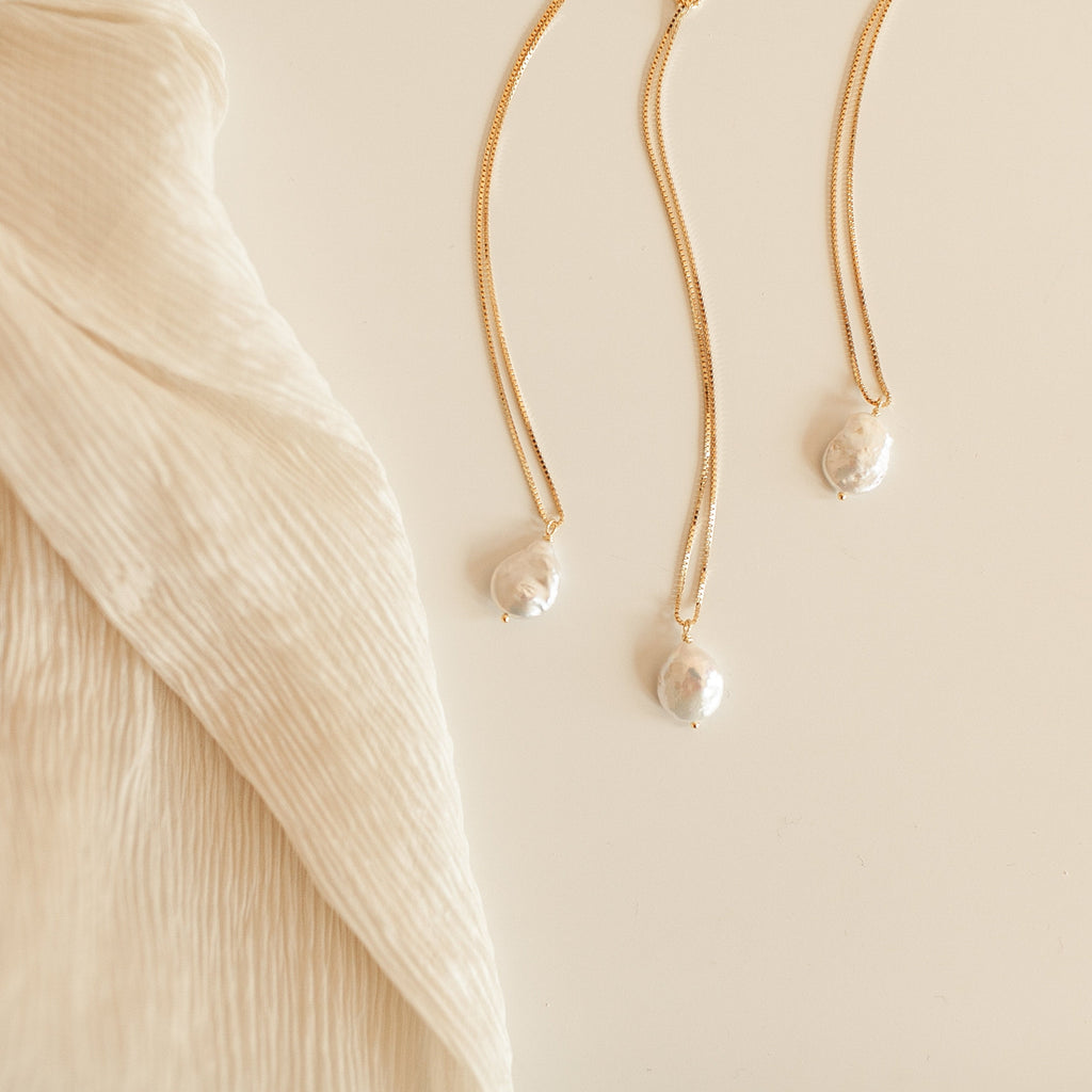 The Sadie Pearl Necklace, featuring a freshwater pearl pendant on a gold chain, rests on soft beige textured fabric—an elegant bridesmaids gift idea.