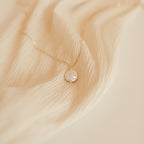 The Sadie Pearl Necklace features a round freshwater pearl pendant on a gold chain, elegantly displayed on beige fabric—an enduring choice for bridesmaids.