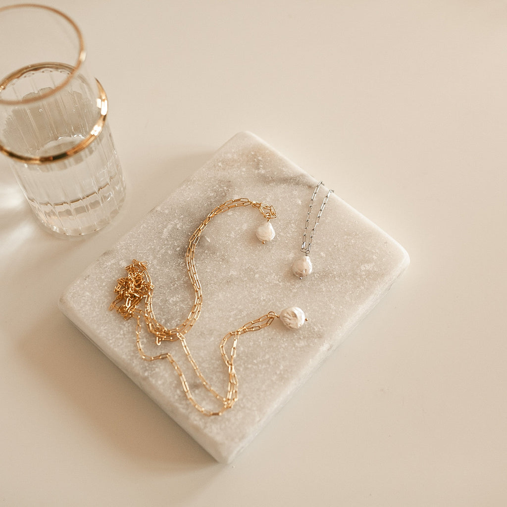 Baroque Pearl Necklaces in gold and silver rest on a marble tray beside a glass with a gold rim.