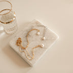 Baroque Pearl Necklaces in gold and silver rest on a marble tray beside a glass with a gold rim.