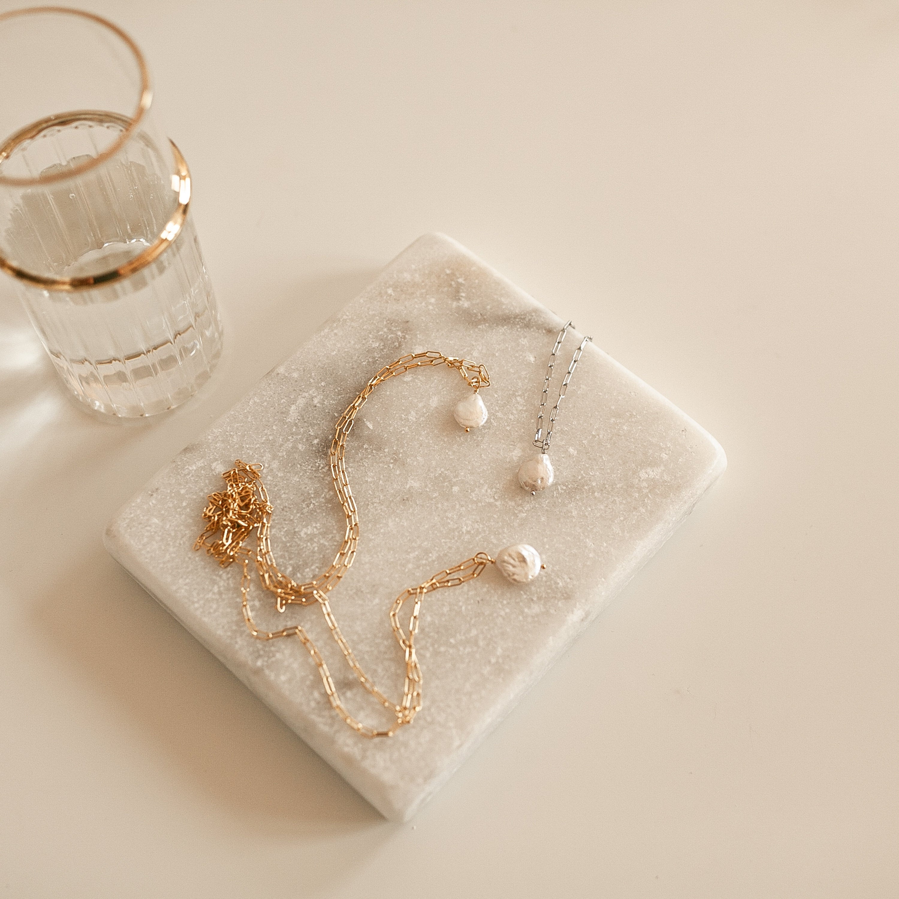 Baroque Pearl Necklaces in gold and silver rest on a marble tray beside a glass with a gold rim.