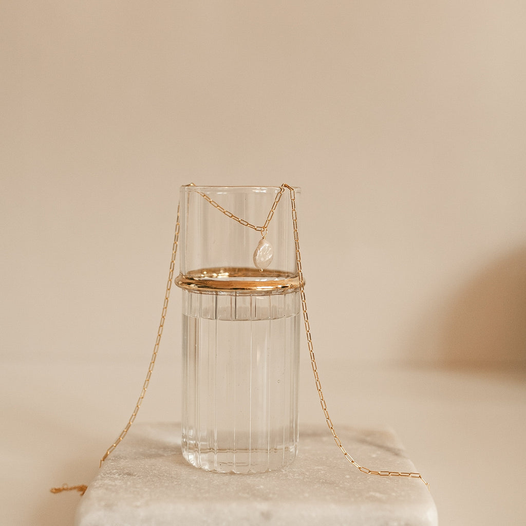 The Baroque Pearl Necklace is elegantly draped over a clear glass of water on a white surface, showcasing its unique pendant.
