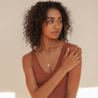 A woman with curly hair in a brown tank top softly touches her shoulder and looks to the side, elegantly showcasing the Baroque Pearl Necklace.