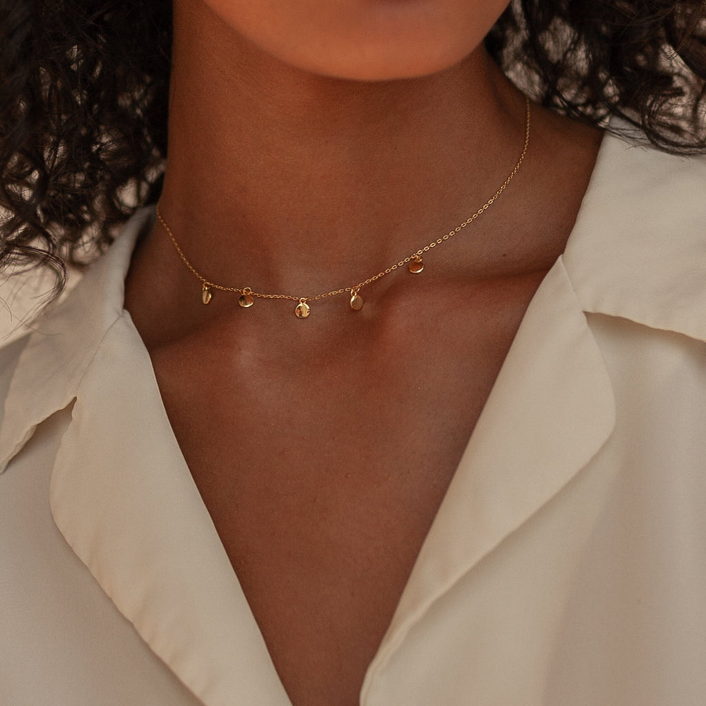 A woman wearing a cream blouse and the Piera Coin Necklace, a delicate gold layering piece with small dangling charms.