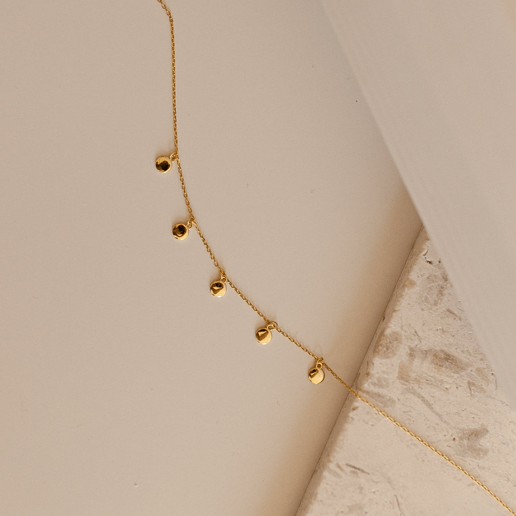 Close up of a delicate gold chain necklace laid on a neutral surface, featuring small polished teardrop charms evenly spaced along the chain.