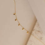 Close up of a delicate gold chain necklace laid on a neutral surface, featuring small polished teardrop charms evenly spaced along the chain.