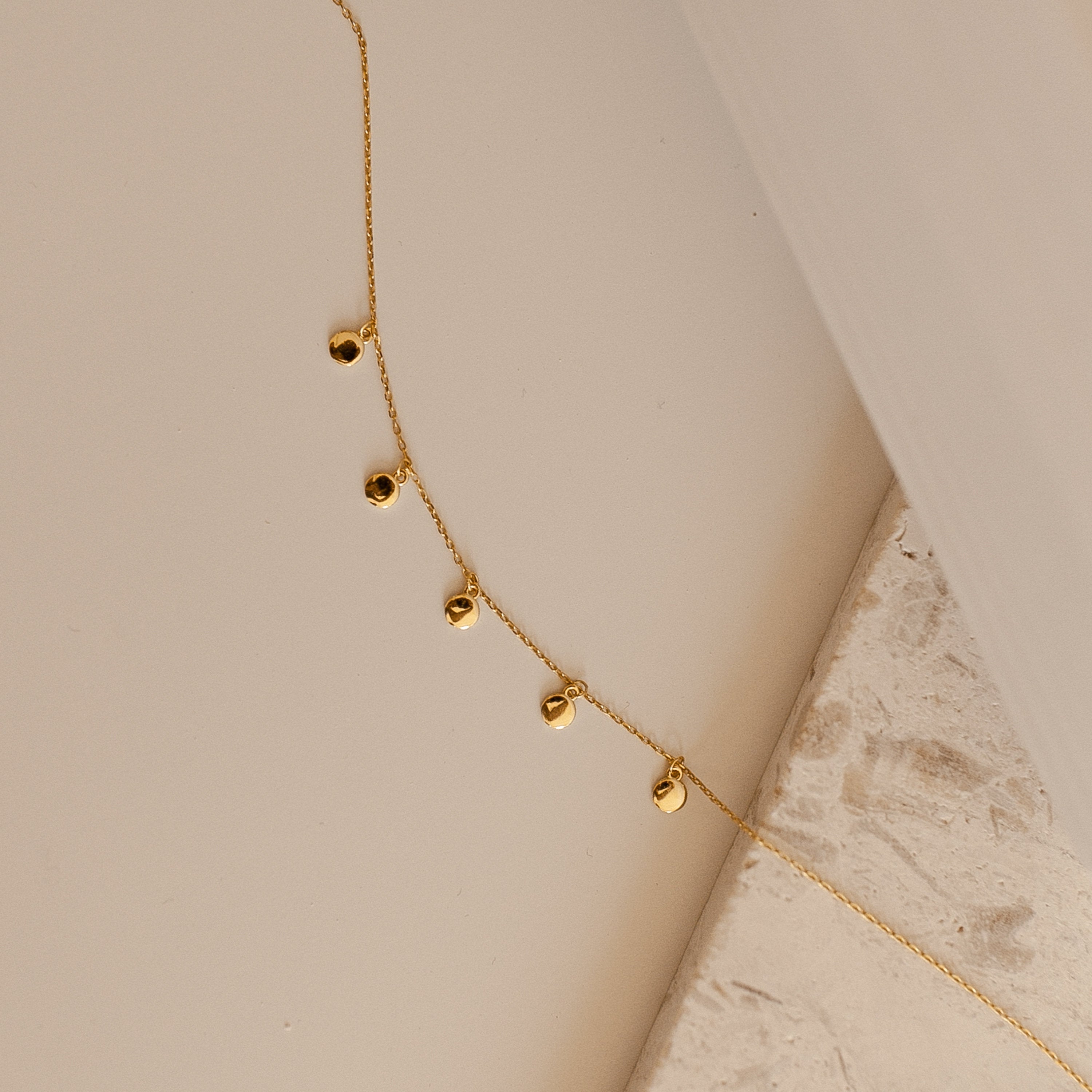 Close up of a delicate gold chain necklace laid on a neutral surface, featuring small polished teardrop charms evenly spaced along the chain.