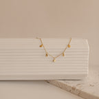 The Piera Coin Necklace, featuring small gold discs, is displayed on a textured white surface with a neutral background—ideal for layering with any outfit.