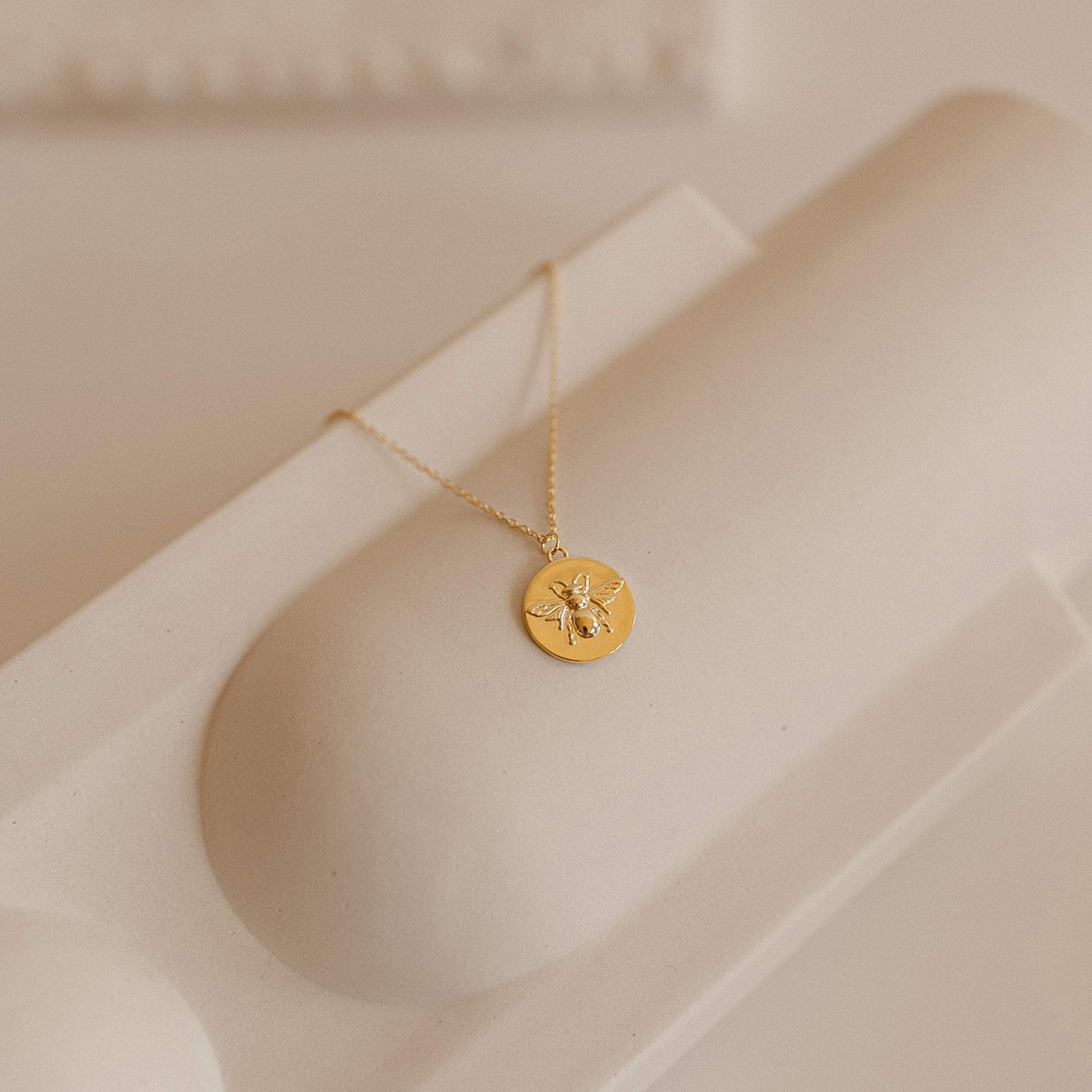 Gold medallion necklace with a raised bee motif, displayed on a curved cream surface. The pendant hangs from a fine gold chain and features intricate wing and body detailing.
