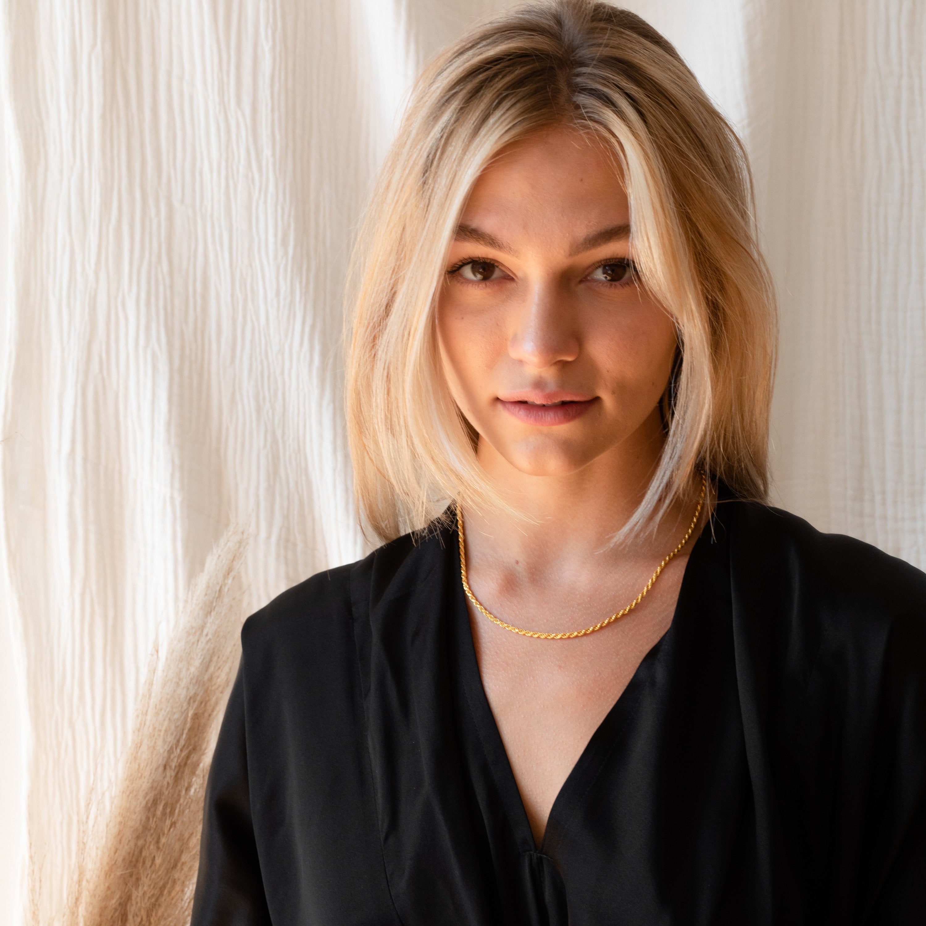 Model with blonde hair wears the gold rope chain necklace with a black robe against a softly lit background.
