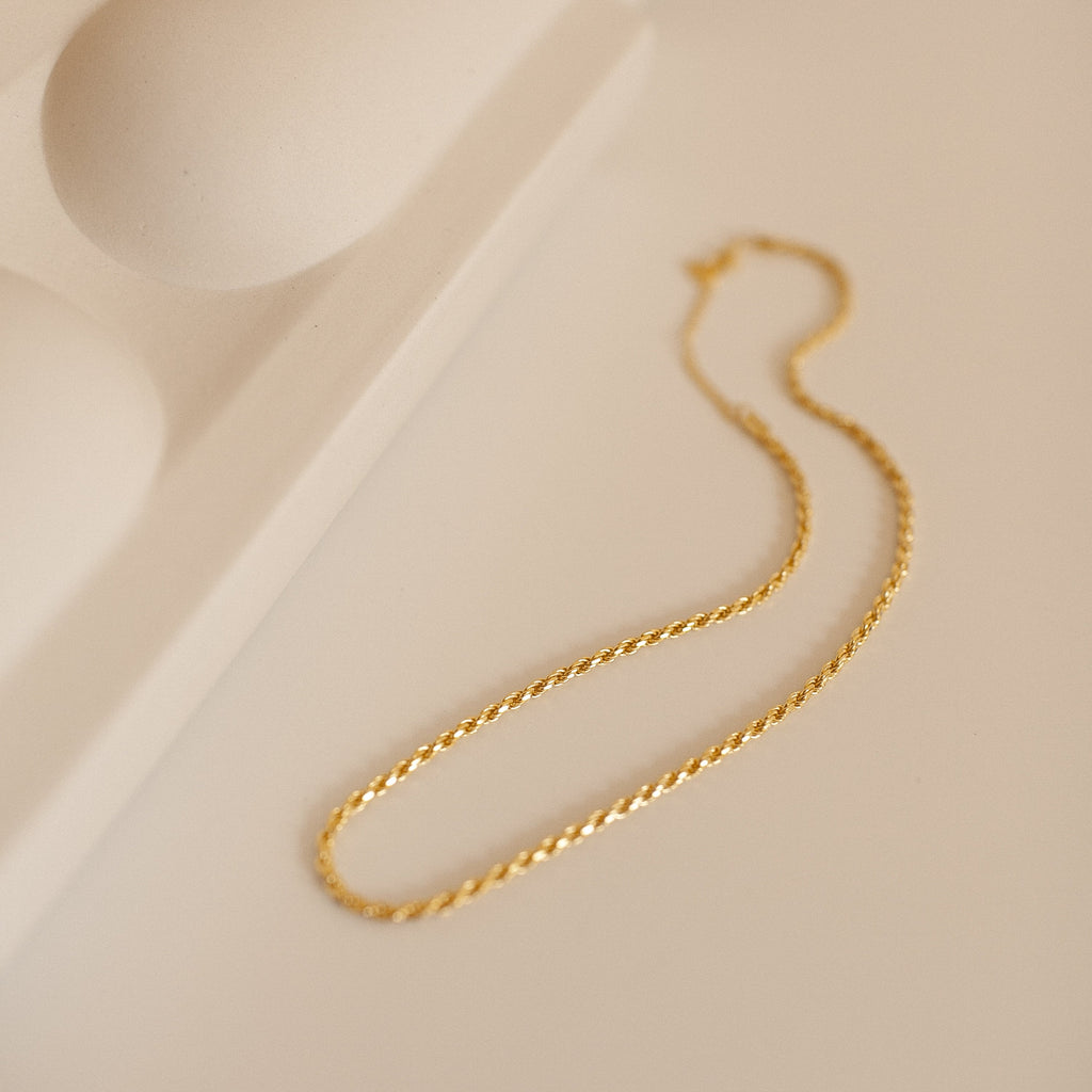 A delicate gold rope chain necklace rests in an elegant S-shape on a soft beige surface beside a sculptural ceramic tray. The chain’s twisted design subtly catches the light, highlighting its fine craftsmanship and intricate texture. 