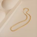 A delicate gold rope chain necklace rests in an elegant S-shape on a soft beige surface beside a sculptural ceramic tray. The chain’s twisted design subtly catches the light, highlighting its fine craftsmanship and intricate texture. 