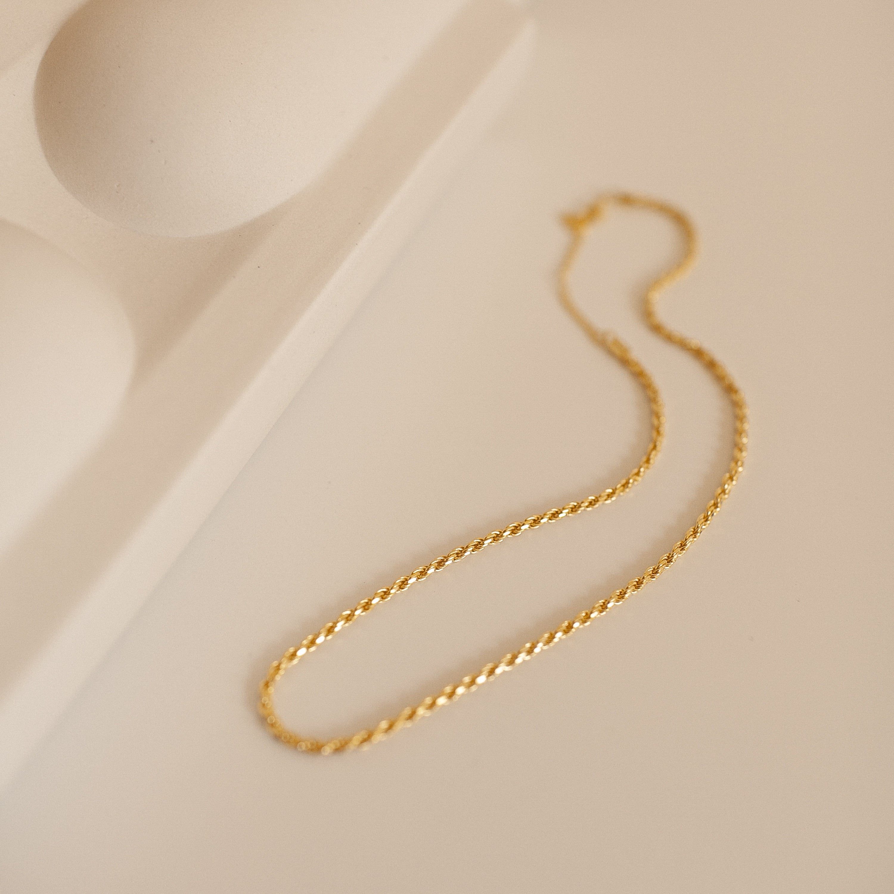 A delicate gold rope chain necklace rests in an elegant S-shape on a soft beige surface beside a sculptural ceramic tray. The chain’s twisted design subtly catches the light, highlighting its fine craftsmanship and intricate texture. 